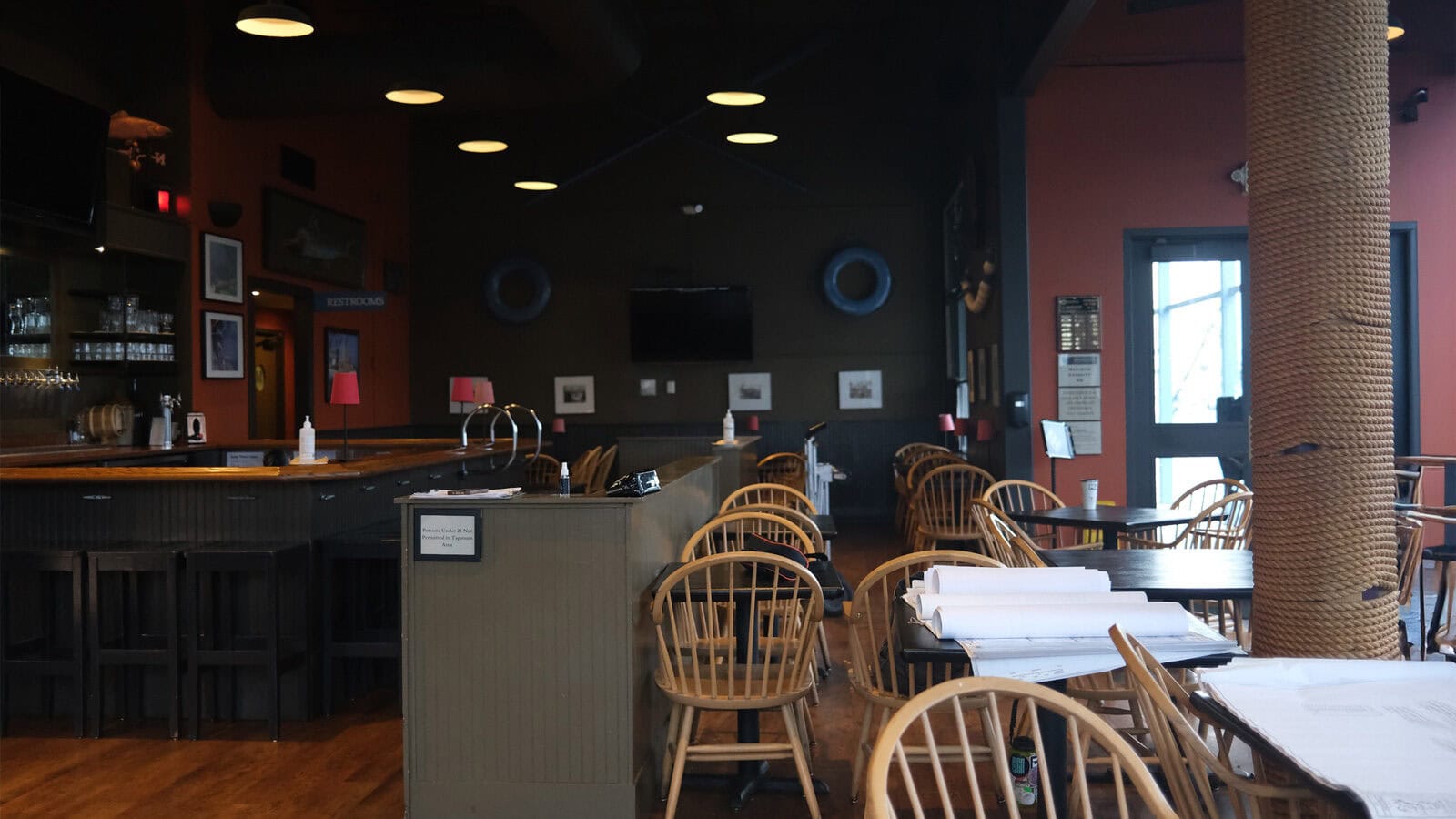The empty Highliner space, soon to become a vibrant Filipino-Hawaiian restaurant, features wooden chairs, tables, and a bar on the left. Its dark walls are adorned with nautical decorations. Dim lighting contrasts with daylight peeking through the window on the right.