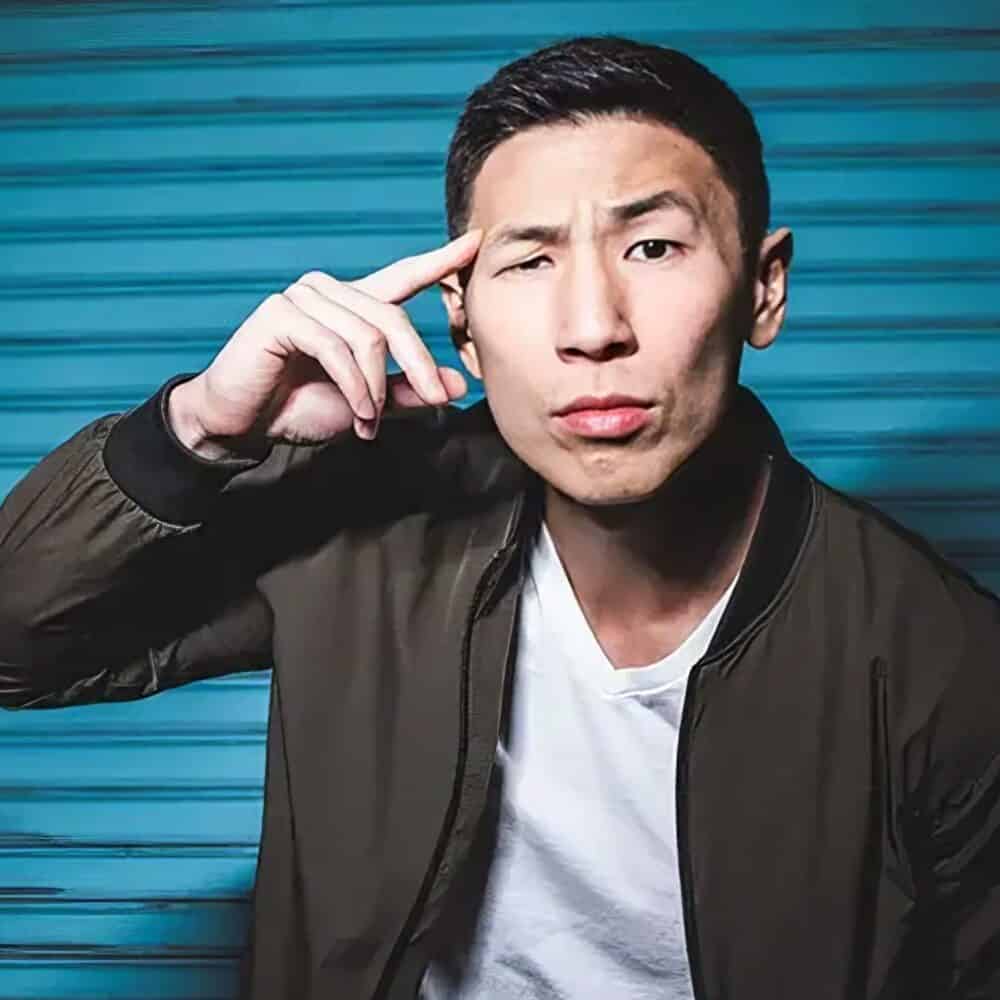 A man in a white shirt and dark jacket stands in front of a blue corrugated background, touching his temple with one hand and looking directly at the camera—an image worthy of any Must List for 05-01-2025.