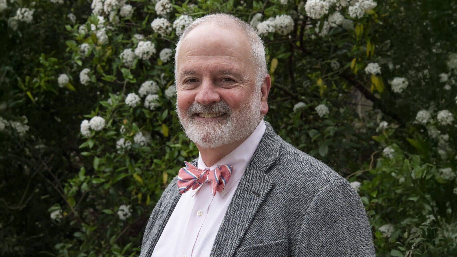 Joshua-Gidding-Headshot_1600x900 An older man with a grey beard, dressed in a grey suit, pink shirt, and striped bow tie, stands before blooming white flowers—like a scene from a book excerpt: old white man writing his thoughts into the spring air.