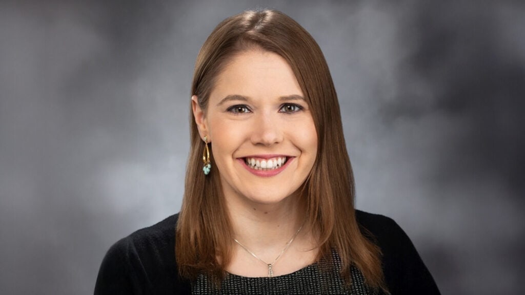 A woman with straight brown hair, wearing a black top, necklace, and earrings, smiles in front of a gray studio background.
