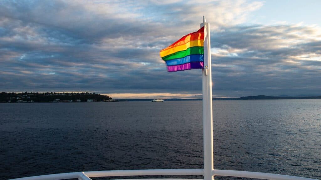 Rainbow flag flying on a boat against a cloudy sky over a body of water. Land is visible in the distance.
