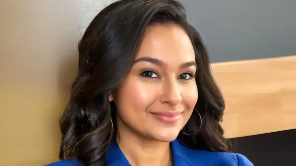 A woman with long dark hair wearing a blue blazer smiles at the camera against a neutral background.