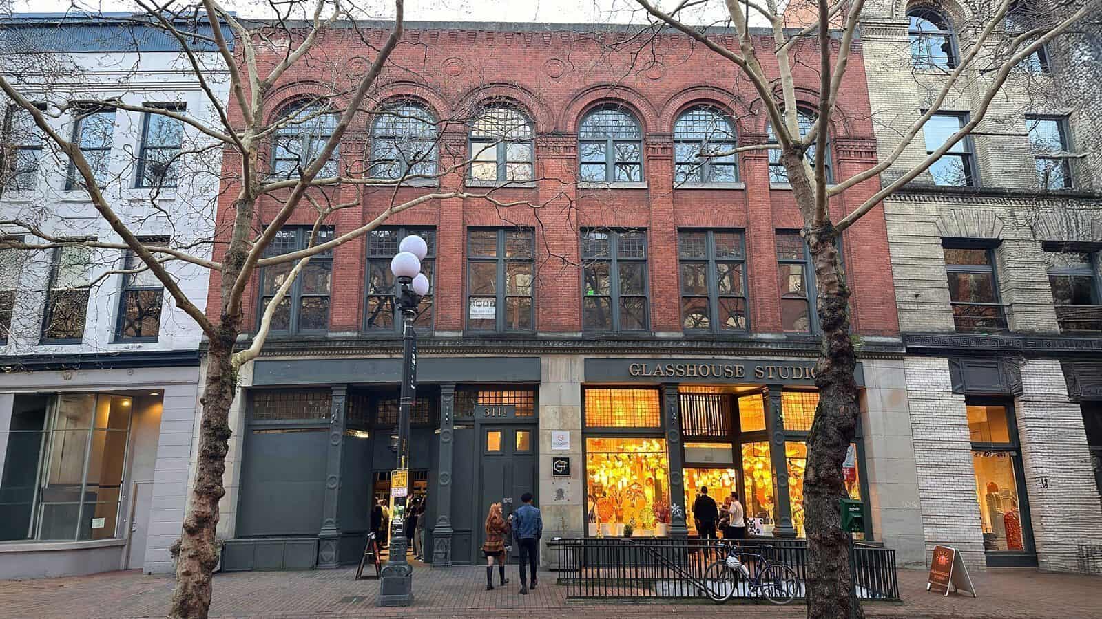Street view of three adjacent buildings with large windows and brick facades. The central one, now opened by former tech entrepreneur Andrew Conru, houses "GLASSHOUSE STUDIO" with lit displays. Bare trees line the sidewalk, celebrating the Seattle Prize Masters Fellowship initiative.