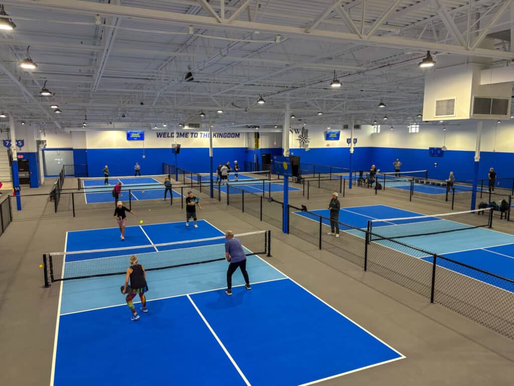 The indoor pickleball facility in Poulsbo boasts multiple blue courts where a lively frenzy of players are either engaged in matches or eagerly waiting their turn. Above, the ceiling's exposed beams and lights add to the dynamic atmosphere.