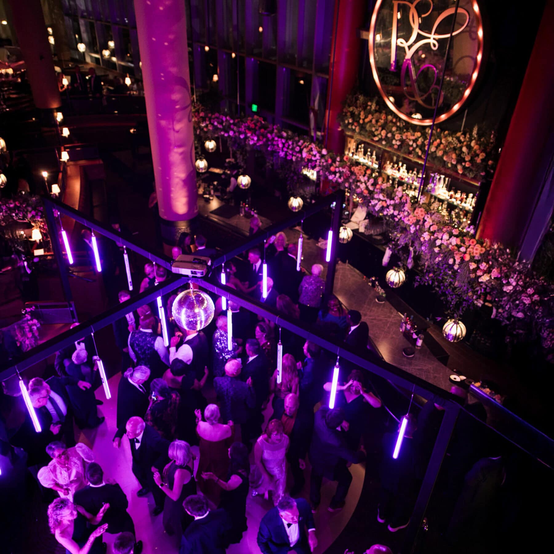 An overhead view of a formal event feels like a modern love story, with people dancing under purple disco lights near a bar decorated with flowers and a large illuminated sign.