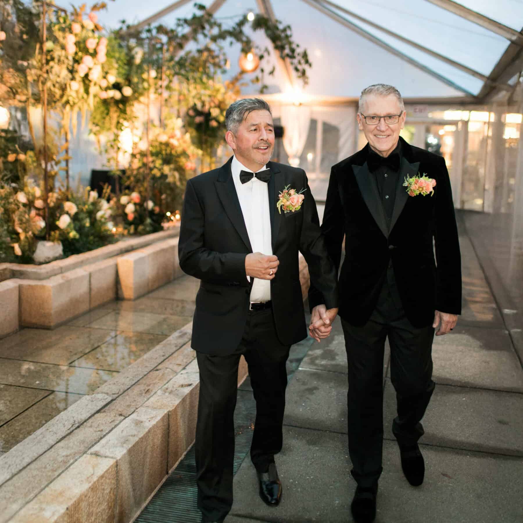 Two men in tuxedos with boutonnieres walk hand-in-hand down an aisle under a clear canopy decorated with flowers and greenery, celebrating their unique love story in this unforgettable moment.