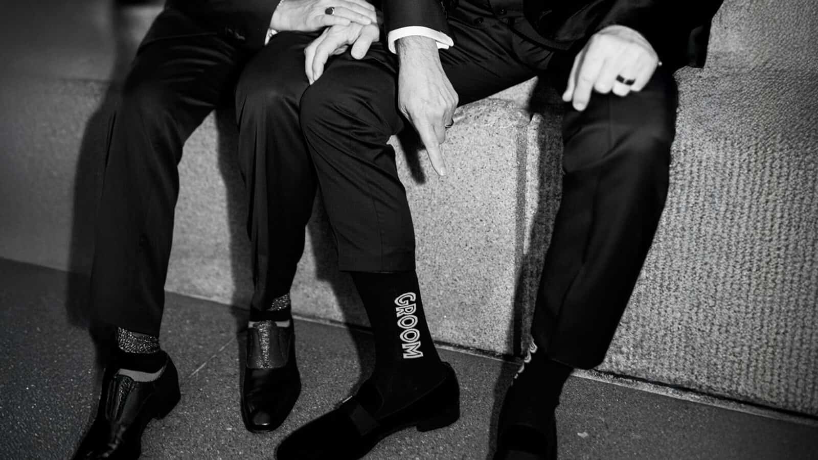 Two people in suits sit side by side; one lifts his pant leg to reveal "GROOM" socks, adding a playful, disco-inspired touch to their love story.