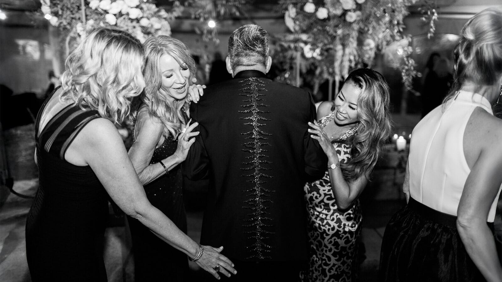 Four women smile and touch the back of a man’s suit jacket at an indoor event, surrounded by floral decorations and disco-inspired vibes.