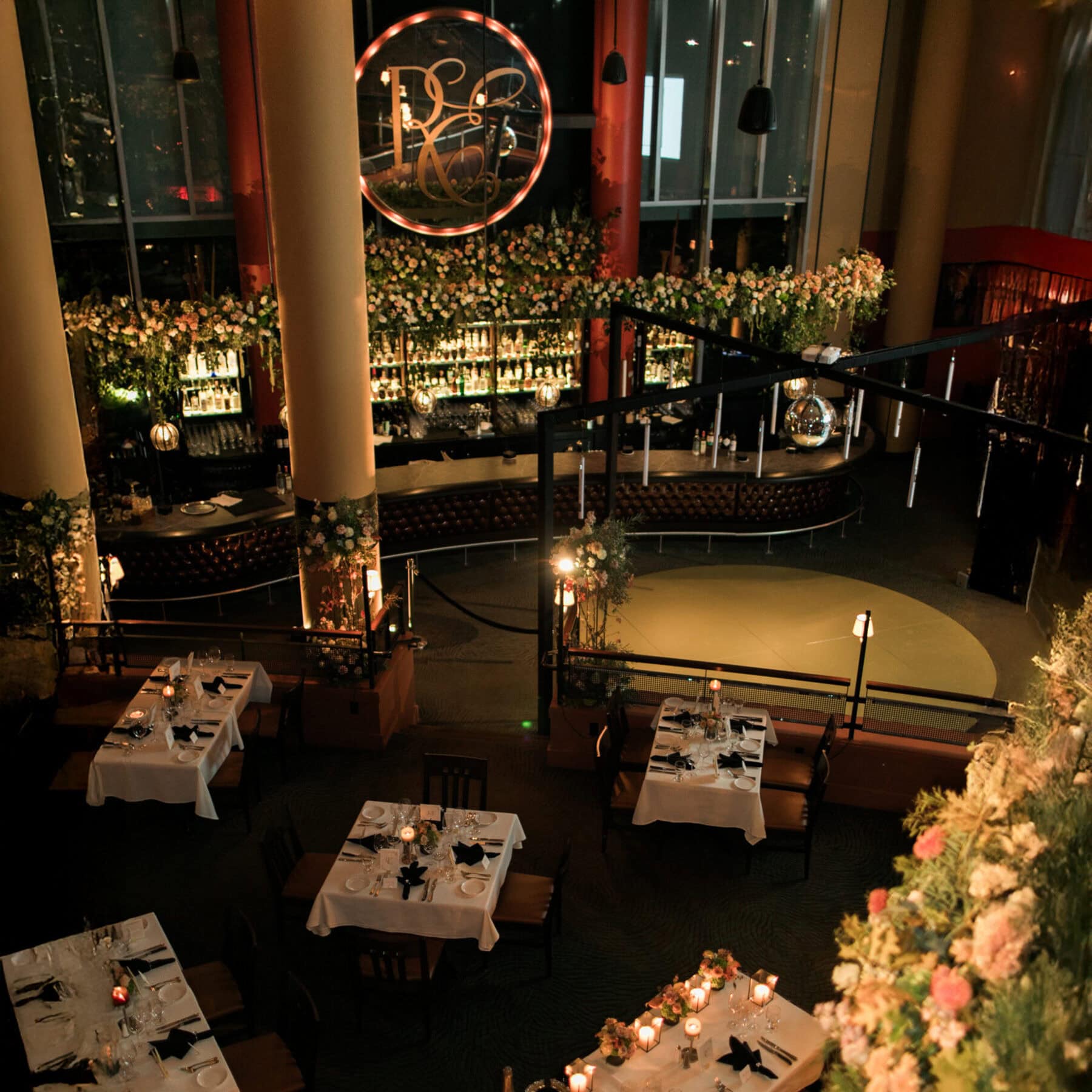 Elegant restaurant interior with white tablecloths, floral decorations, a large illuminated wall sign, and a well-stocked bar in the background. Evening lighting and subtle disco-inspired touches create a warm, inviting ambiance.