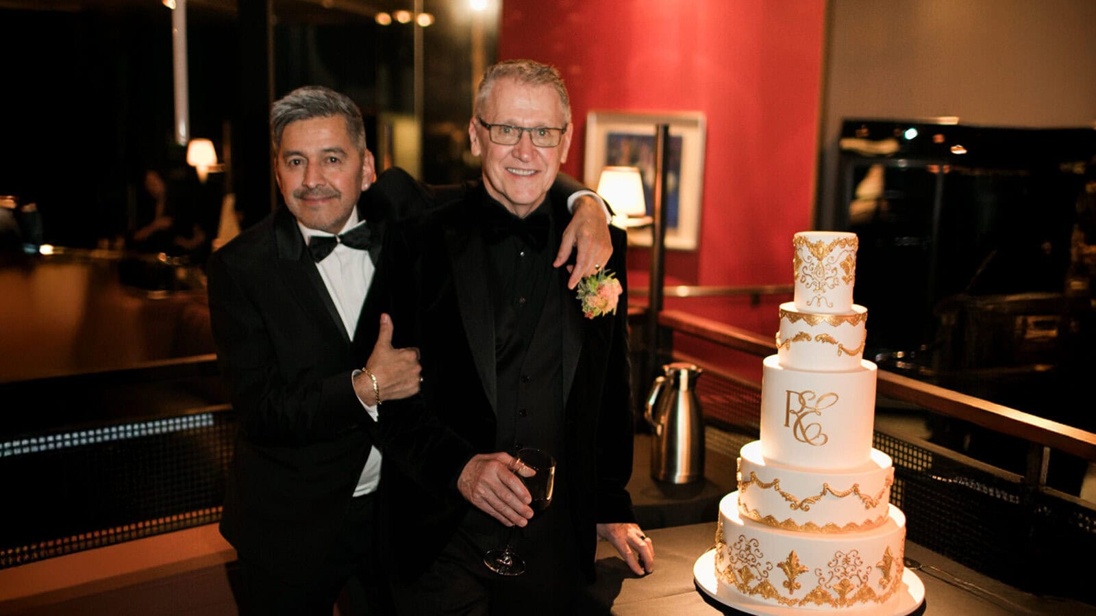 Two men in formal attire stand beside a multi-tiered white and gold cake; one man holds a wine glass, and both are smiling at the camera, celebrating their love story with disco-inspired flair.