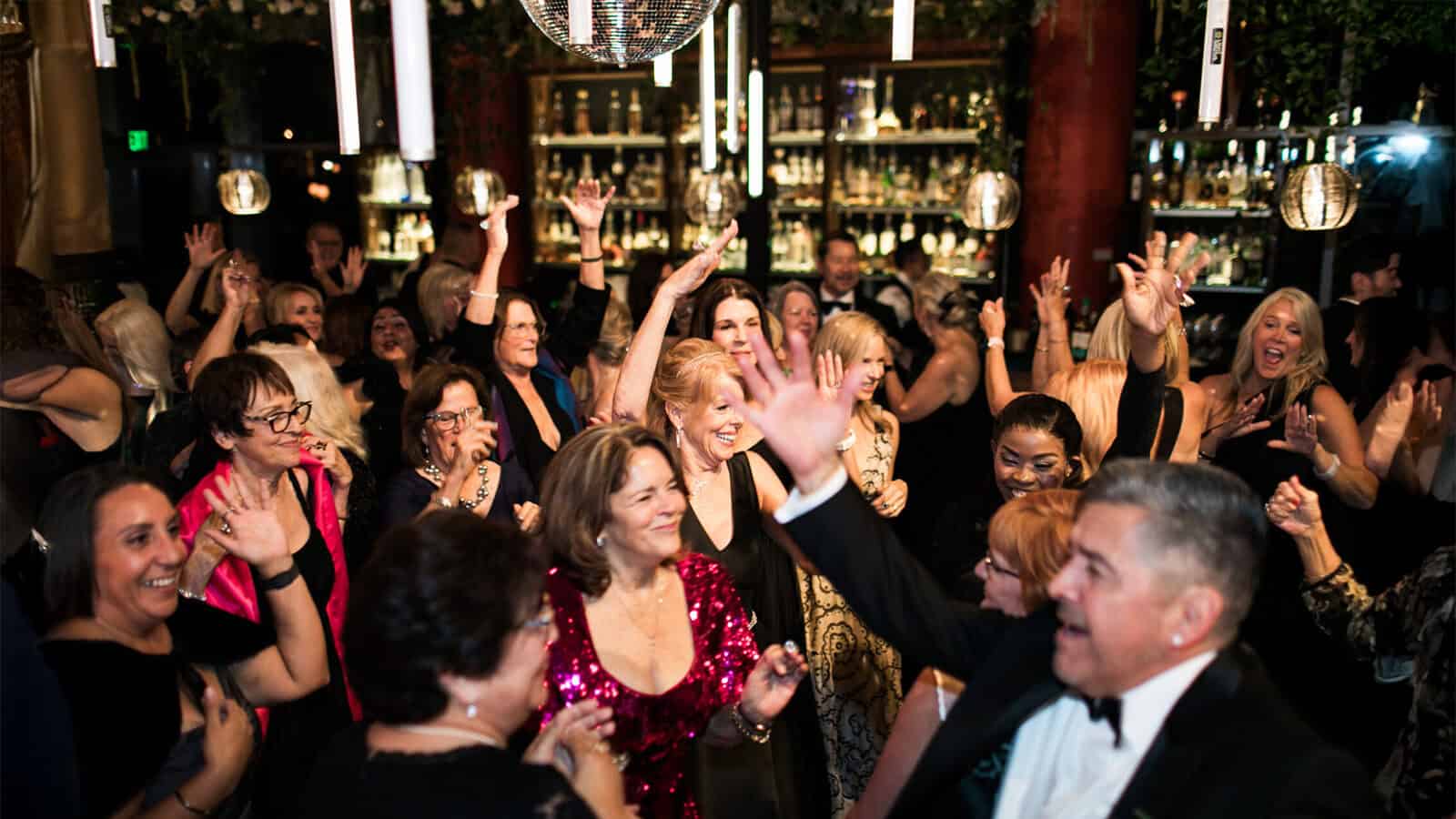 A large group of people dressed formally are dancing and celebrating together at an indoor party with festive lighting, decorations, and a disco-inspired vibe.