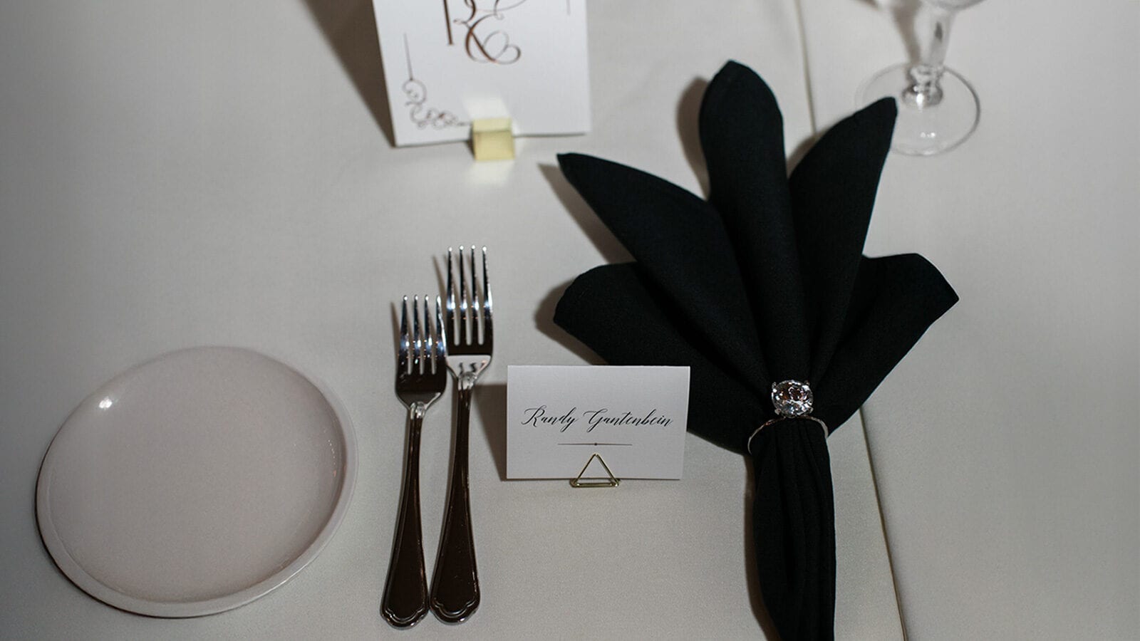 A formal table setting with a white plate, two forks, a name card reading "Rocky Gonzalesian," and a folded black napkin with a napkin ring brings disco-inspired charm to the scene, complemented by a wine glass on a crisp white tablecloth.