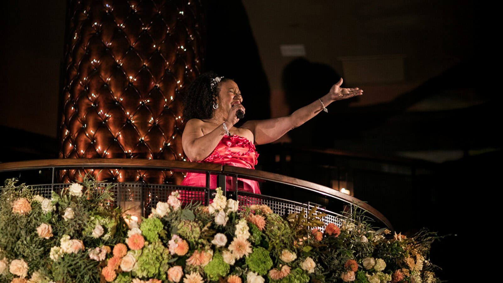 A woman in a red dress sings into a microphone on a flower-adorned balcony during an indoor event, creating a dazzling disco-inspired atmosphere.