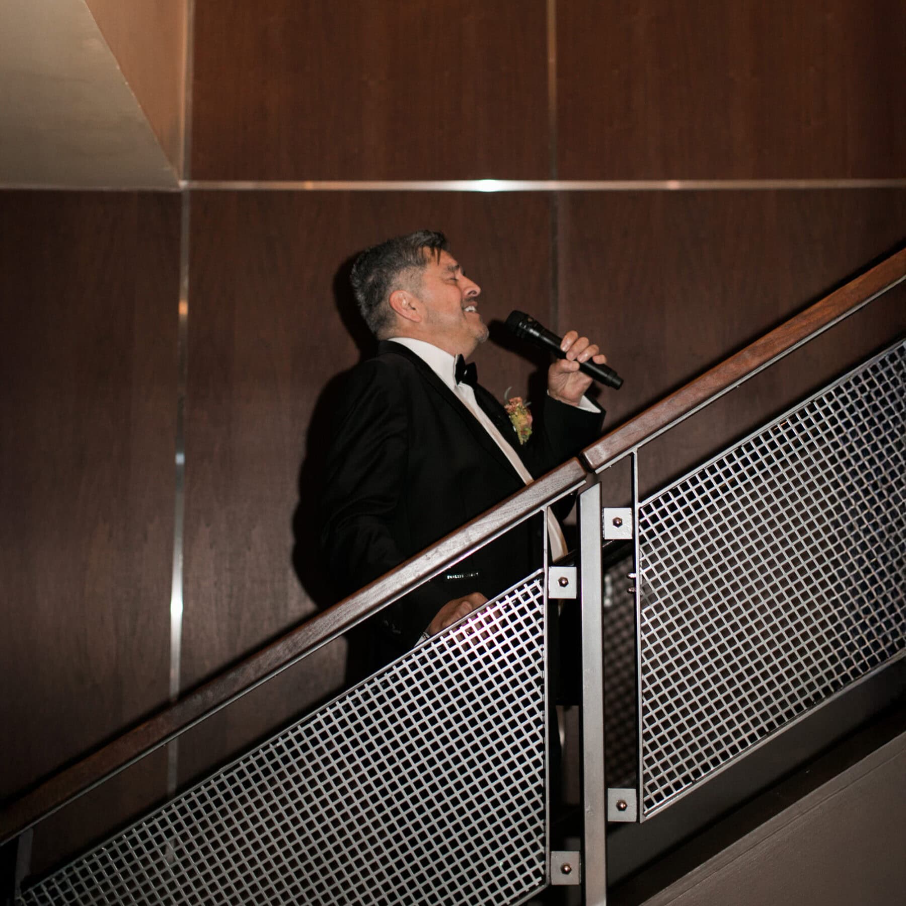 A man in a suit stands on a staircase, holding a microphone and speaking, with a dark wood-paneled wall in the background, evoking a disco-inspired atmosphere.