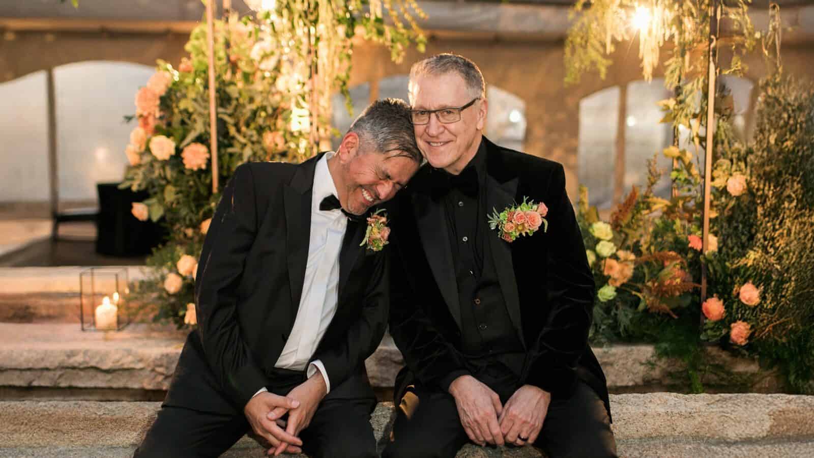 Randy-Ernie-Wedding_1600x900 Two men in tuxedos, with floral boutonnieres, sit closely on a stone bench amid floral decorations, appearing content and relaxed at an event.