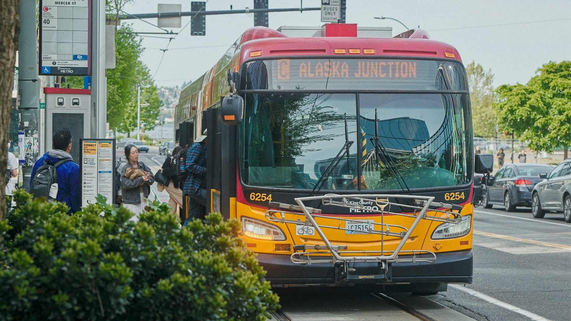 A Seattle Metro bus labeled "C Alaska Junction" stops to pick up passengers at a bus stop on a city street.