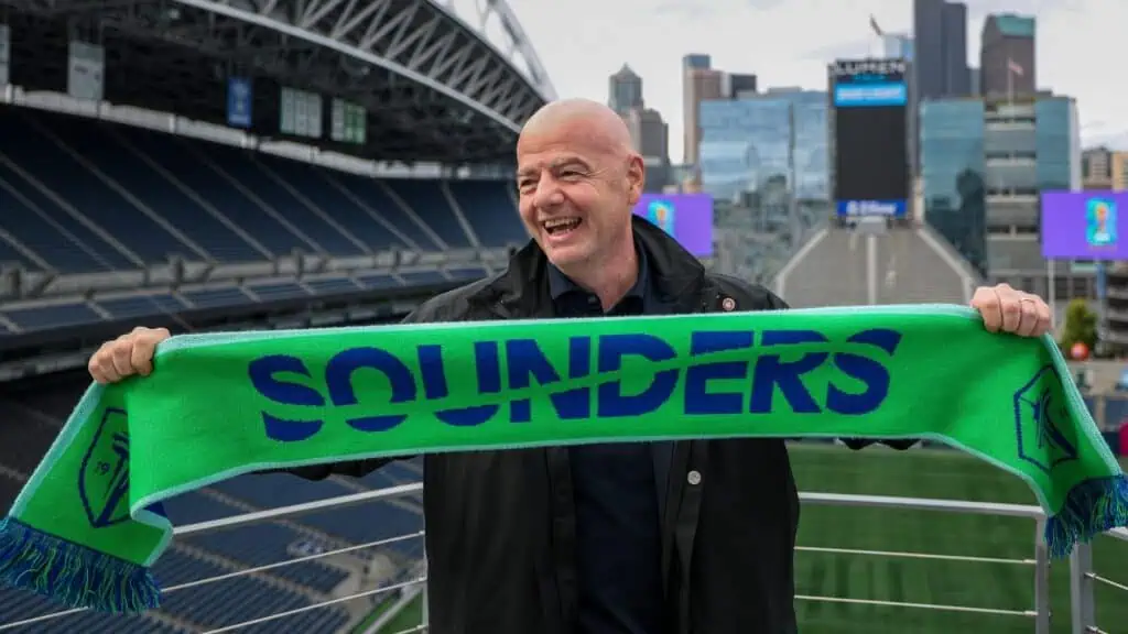 A man stands in a stadium among soccer fans, holding a green and blue scarf with the word "SOUNDERS" on it, city buildings visible in the background.
