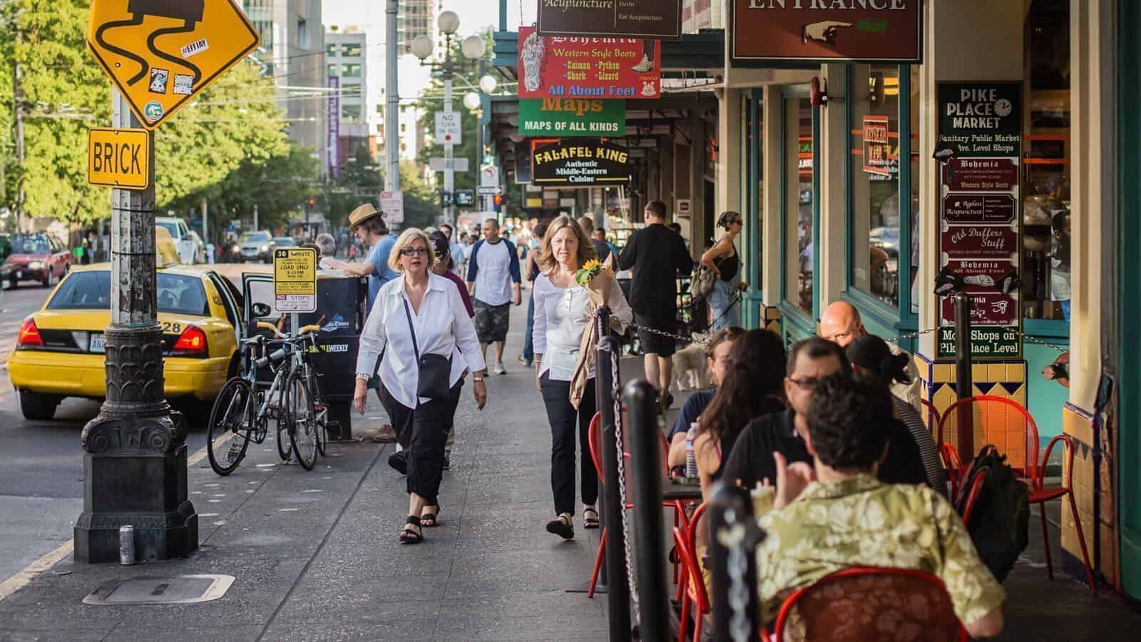 Seattle-CentralBusinessDistrict_1600x900 People walking on a busy sidewalk lined with shops and outdoor diners, near a street with cars and bicycles.
