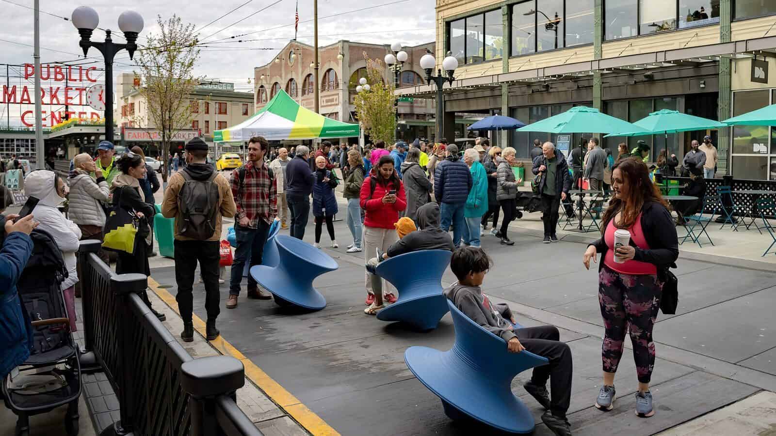 Seattle-First-And-PikeSt_1600x900 In this lively street scene, The Changing Face of First and Pike unfolds. People mingle at the market, some perched on blue spinning chairs while others exchange pleasantries. Behind them, a building adorned with green umbrellas stands as a backdrop to the vibrant interactions.