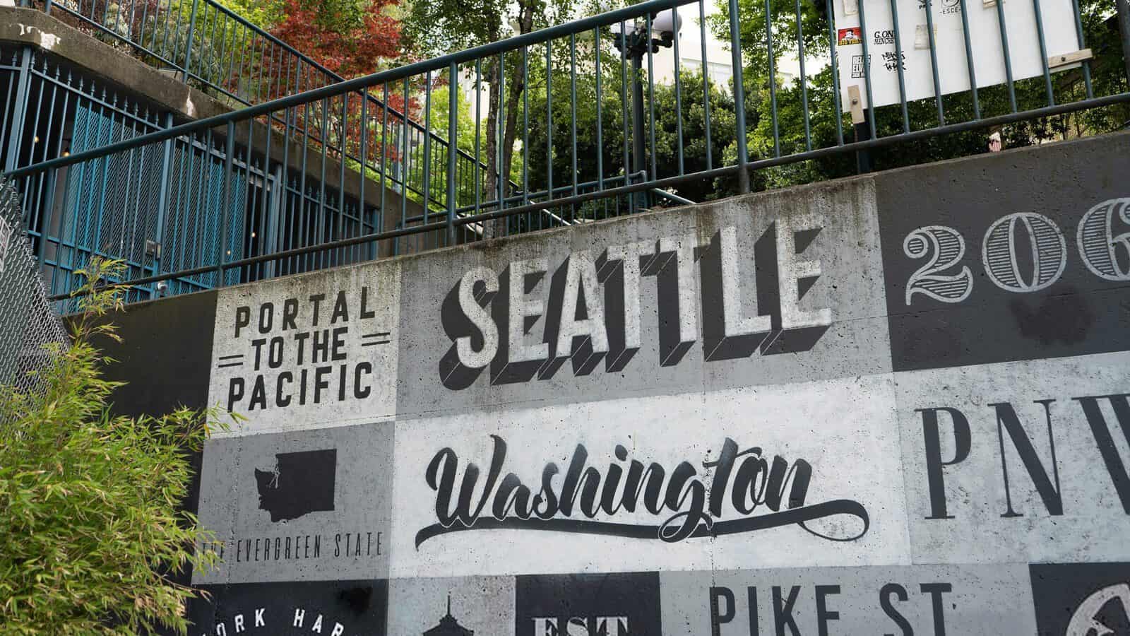 SeattleUrbanGraffiti_1600x900 Concrete wall adorned with phrases like "Seattle," "Washington," and "Portal to the Pacific" in various fonts, proudly marking the City of Cool. Above, a fence and greenery complete this vibrant scene.