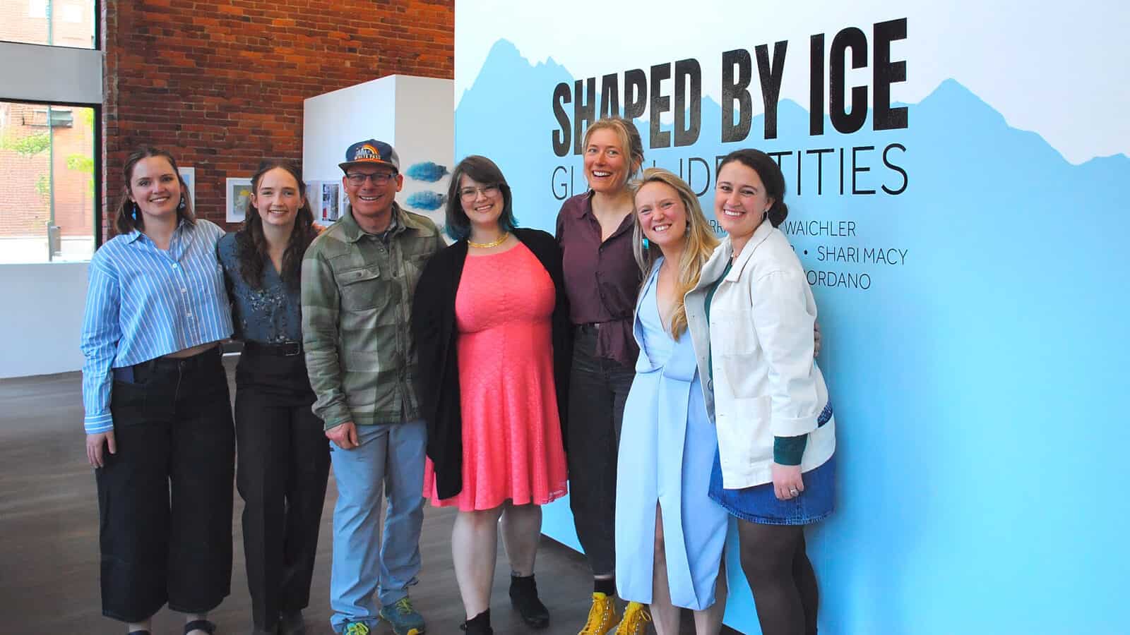 Seven people stand smiling in front of a blue and white wall with the text "Shaped by Ice: Glacial Identities," showcasing their unique Glacial Expressions at an indoor gallery or exhibition space.