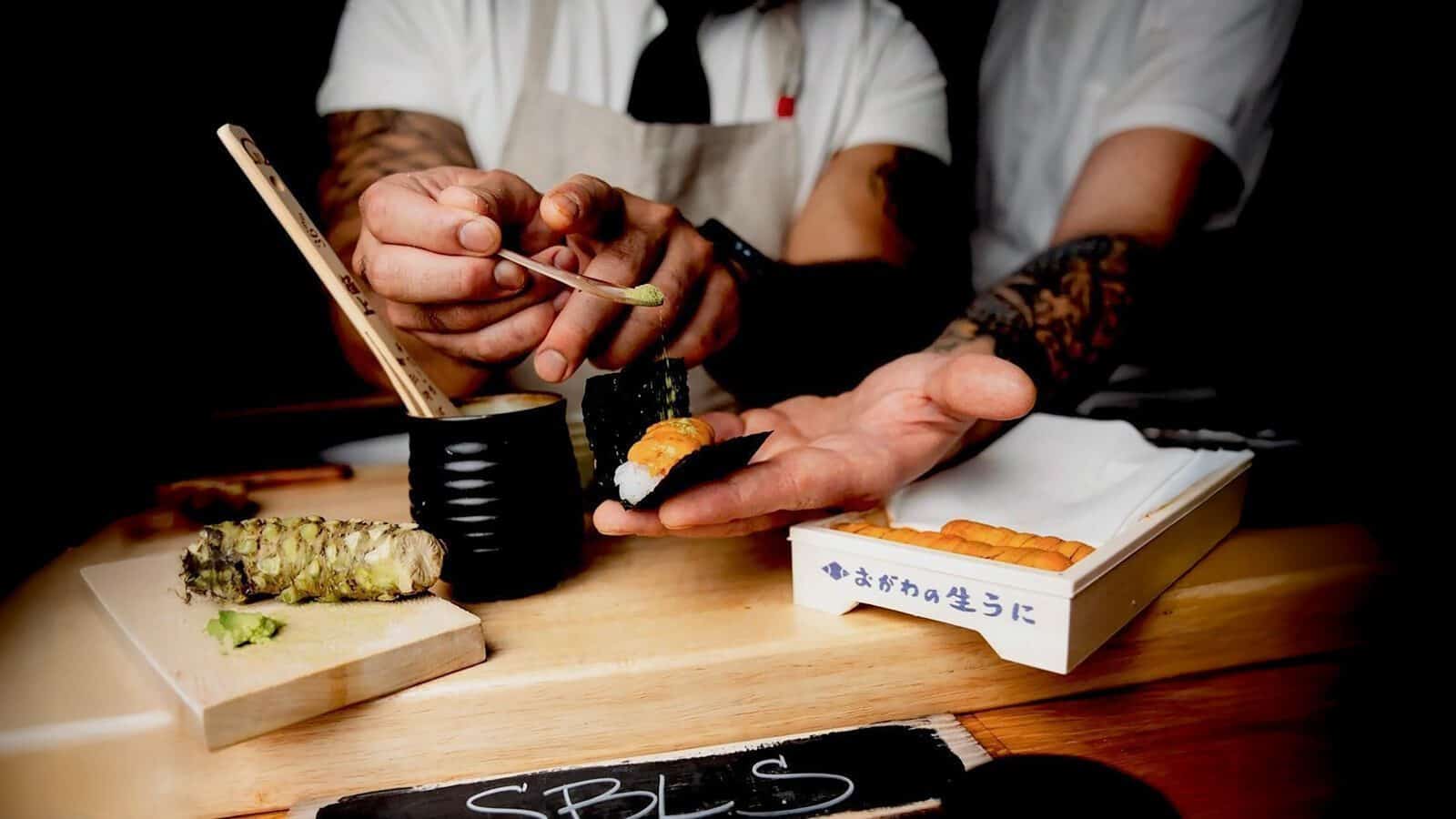 SushiByScratch_1600x900 Chef placing wasabi on sushi while another person holds it, capturing a moment worthy of the latest food news. A whole wasabi root and box of sushi sit enticingly on the counter, adding to the vibrant culinary scene reminiscent of a Restaurant Roundup.