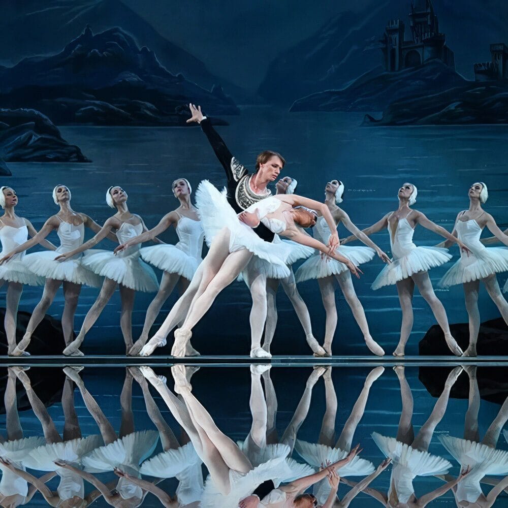 Ballet dancers perform on stage in white costumes, with a male dancer supporting a female dancer in a dramatic pose. A painted lake and castle set is in the background—a must list event for 04-24-2025.