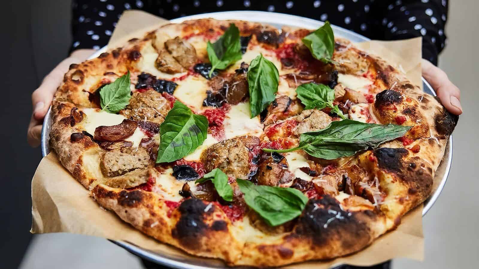 TheLocal104-Pizza_1600x900 A person holds a round pizza on parchment paper, topped with basil leaves, sausage, cheese, and tomato sauce on a slightly charred crust—just one of the mouthwatering creations Hidden in Plain Sight at The Local 104.