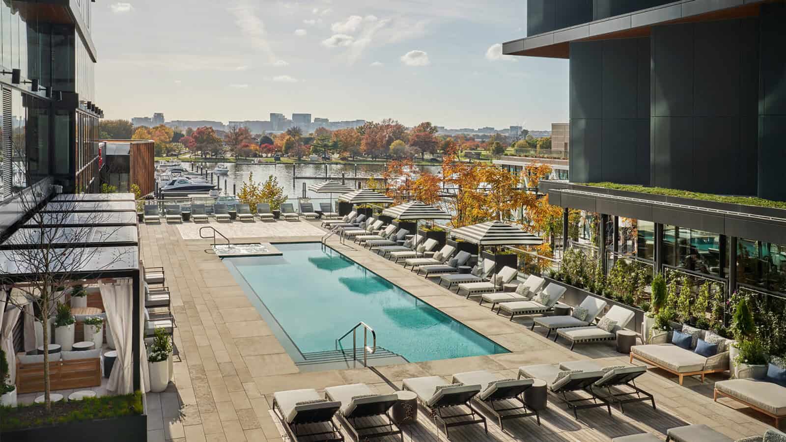 Modern outdoor pool area with lounge chairs, cabanas, and umbrellas, overlook a marina framed by autumn trees and city buildings—a perfect setting for A Stroll Through Old & New D.C. beneath a partly cloudy sky.