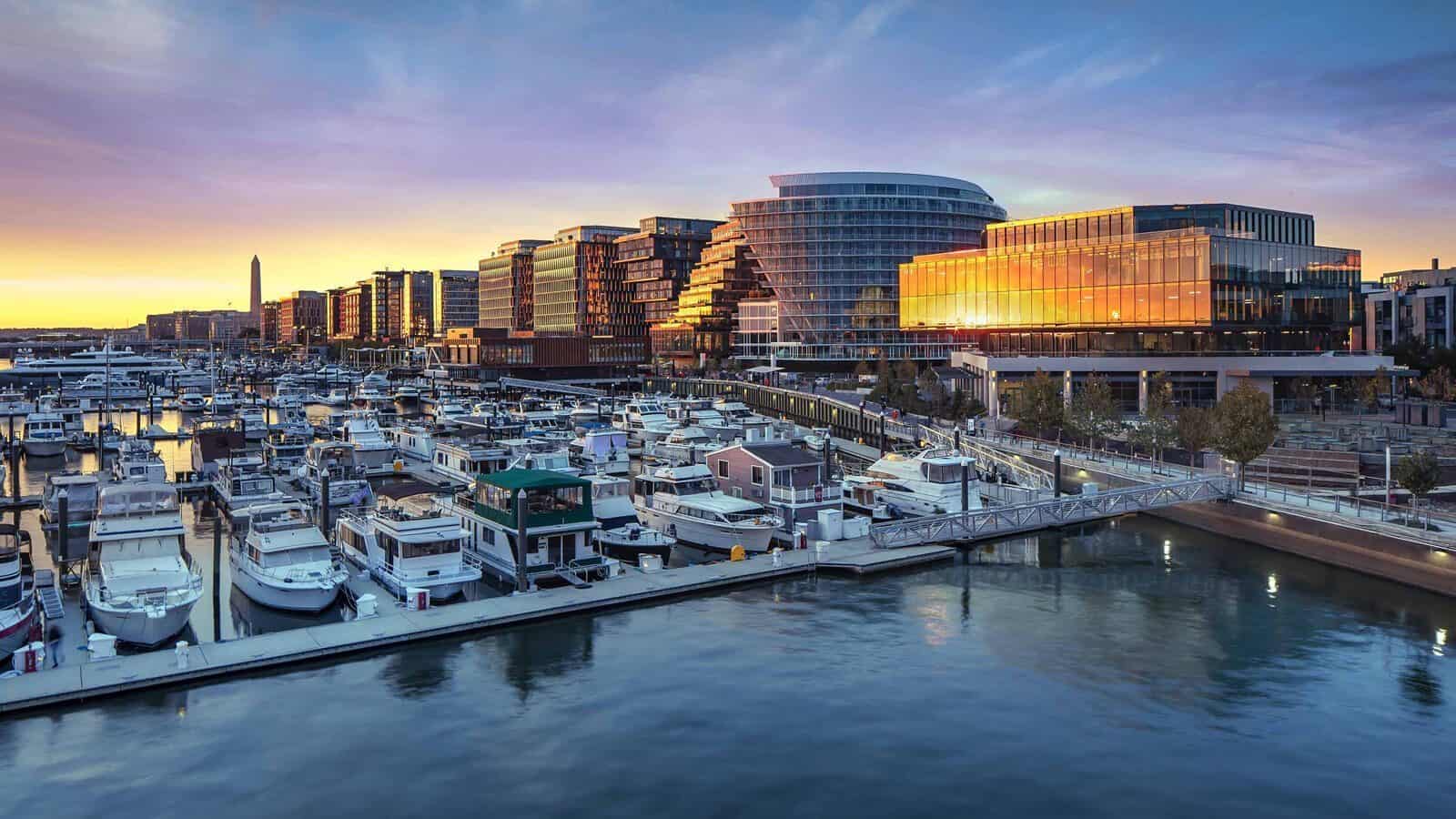 TheWharf-DistrictWharf_1600x900 Modern waterfront buildings and a marina with docked boats at sunset reflect light on the water—capturing the essence of A Stroll Through Old & New D.C. against an urban skyline in the background.