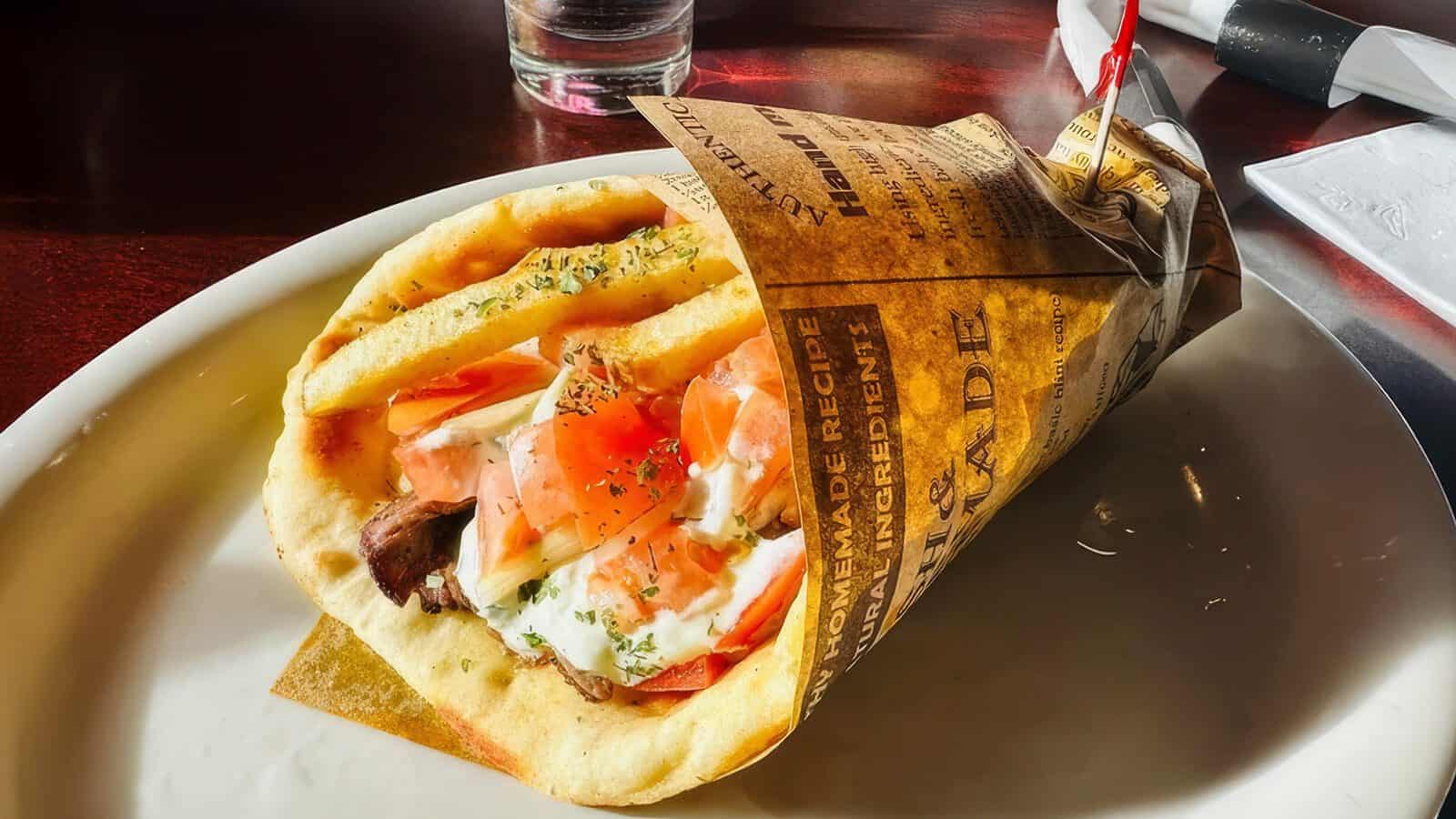 TimeOutRestaurant-Gyro_1600x900 A comfort food classic, this gyro wrap is filled with sliced meat, tomatoes, onions, and tzatziki sauce, served on a white plate and partially wrapped in newspaper-style paper.