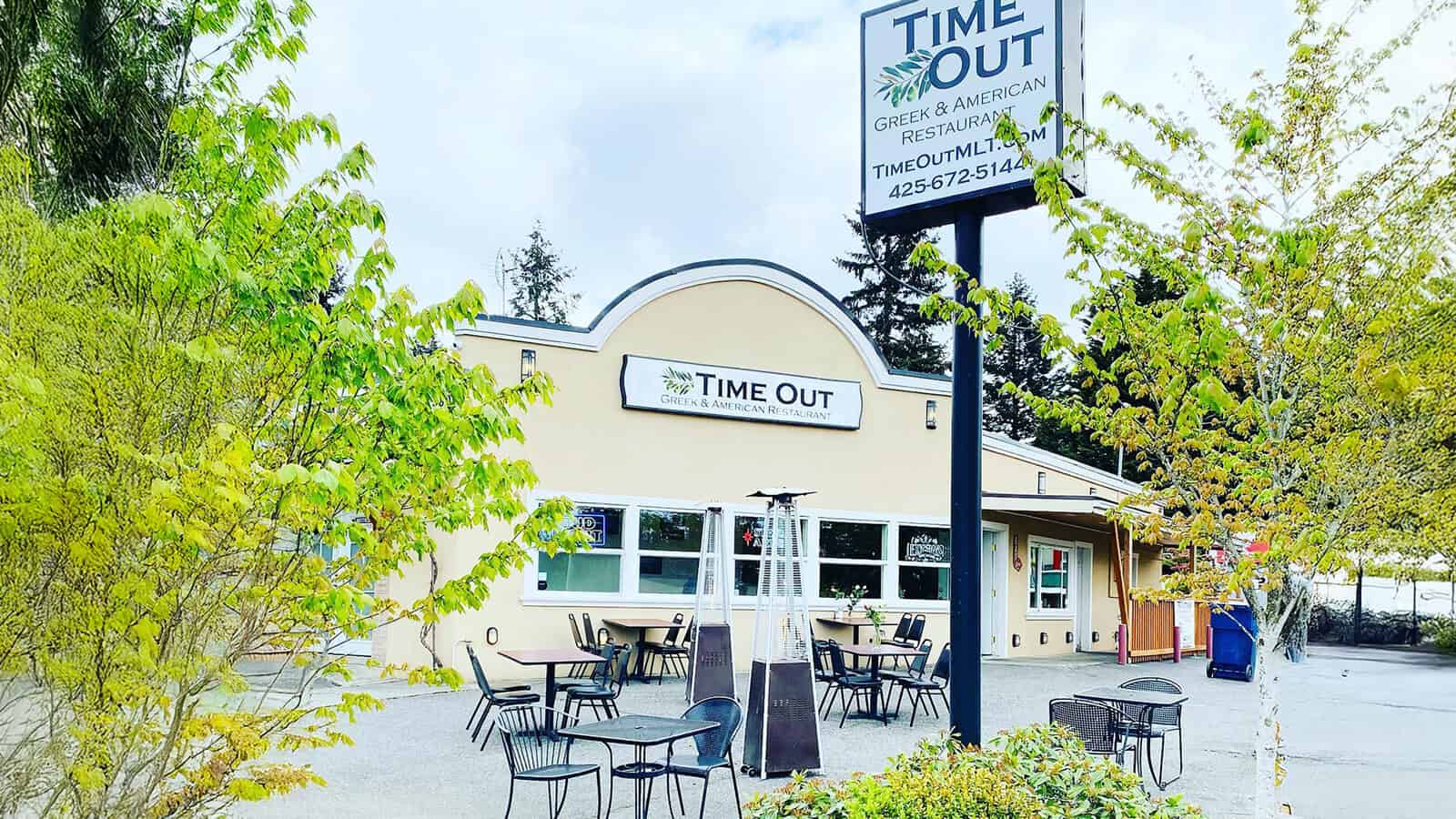 Outdoor seating area with several tables and chairs in front of the Time Out Greek & American Restaurant, perfect for enjoying comfort food like a classic Time Out Burger on a partly cloudy day.
