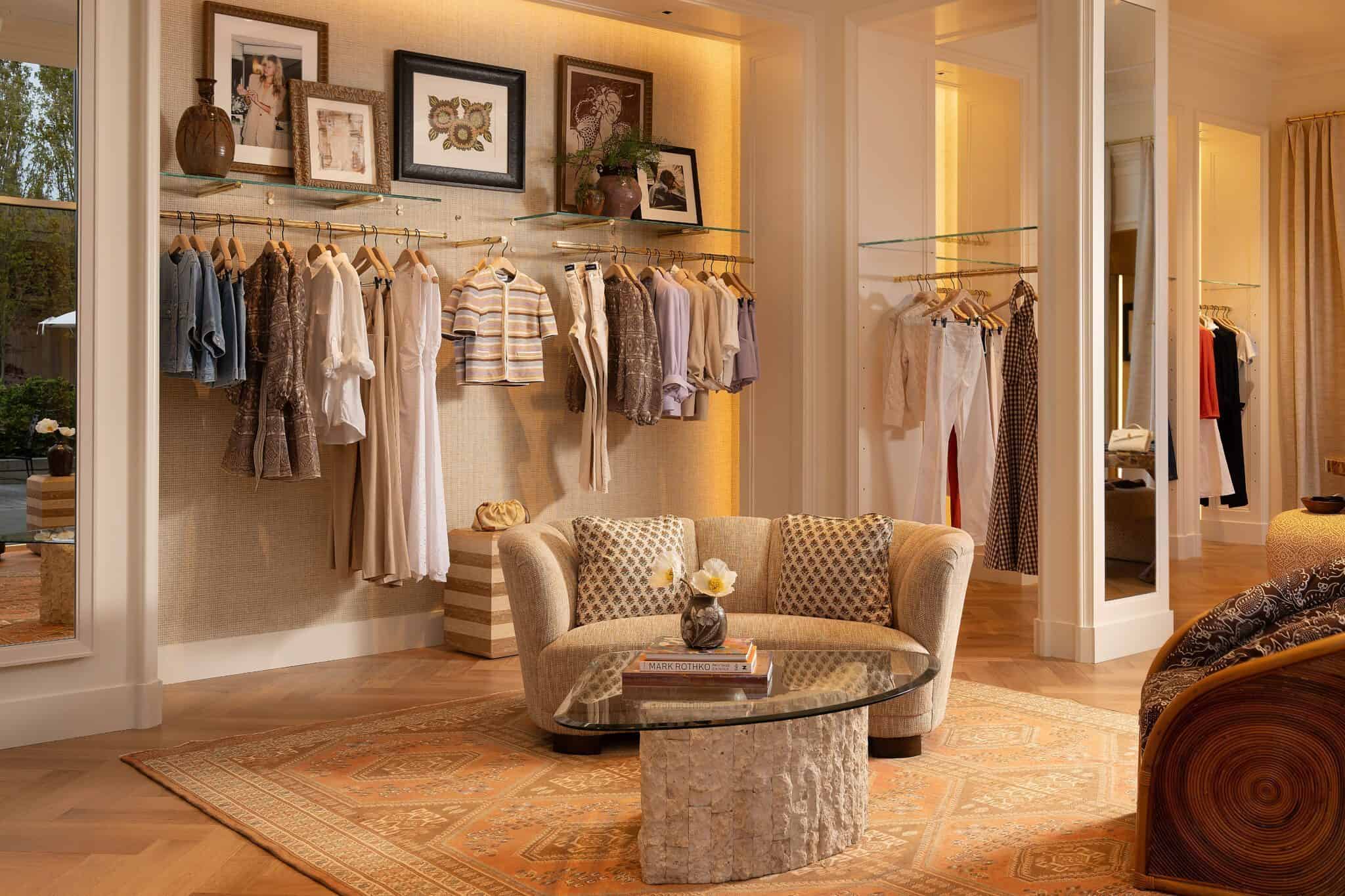 VERONICA BEARD A cozy room with a beige couch, glass coffee table, and a display of hanging clothes. Art and a plant decorate the wall. A warm rug covers the wooden floor.