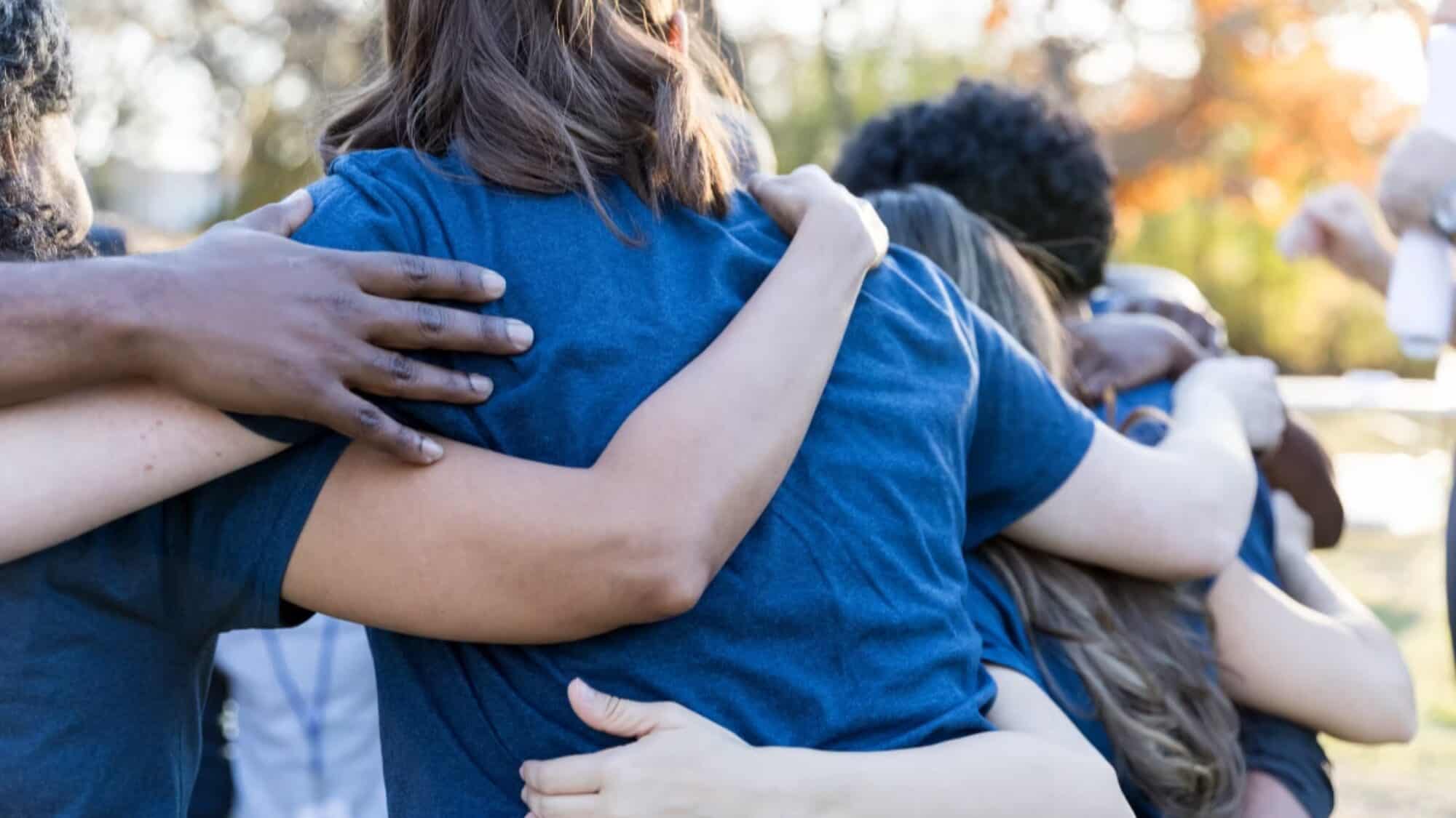 A group of people wearing blue shirts stand outdoors with their arms around each other, embodying the spirit of camaraderie fostered by Eastside Youth Services.