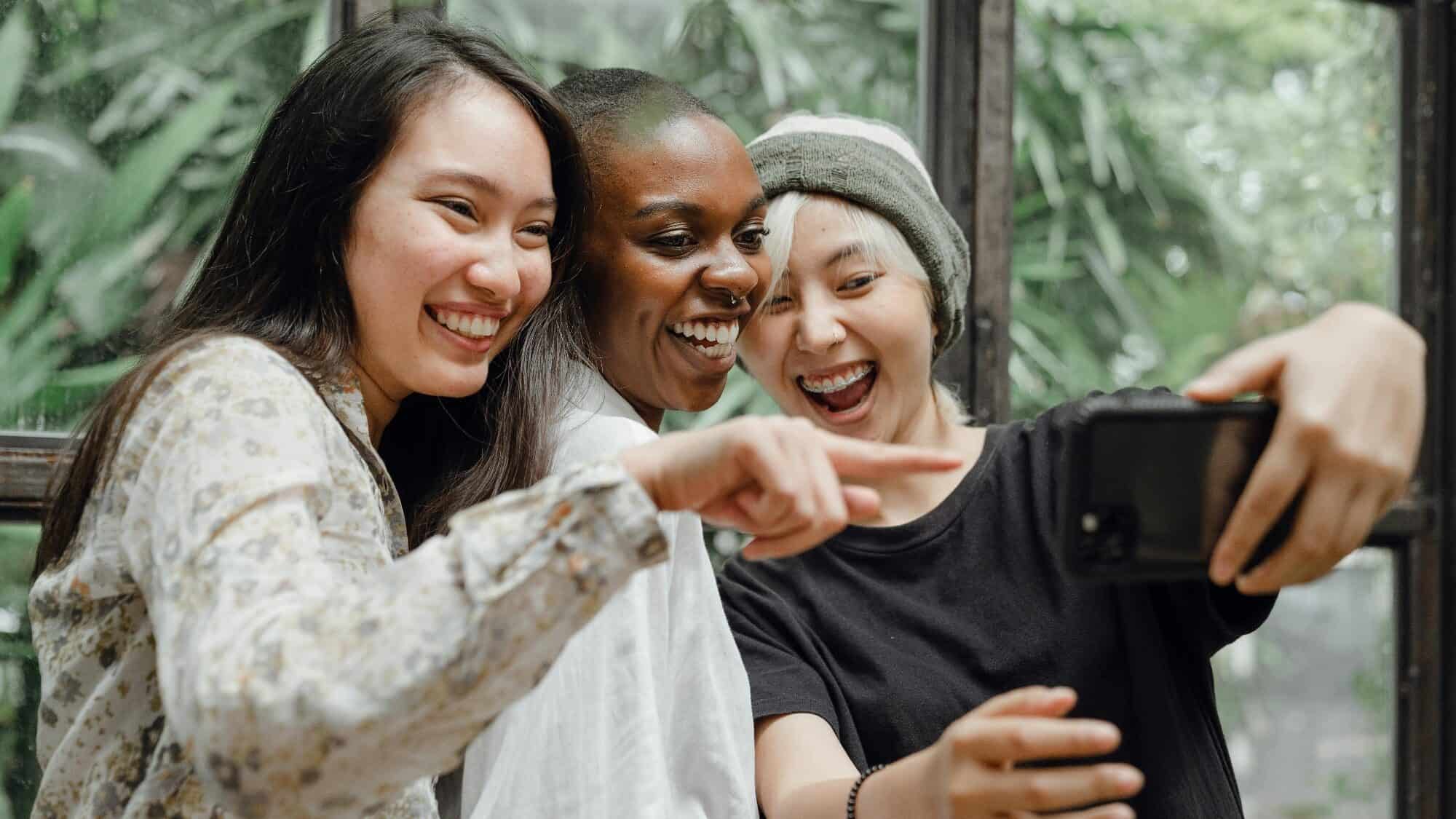 pexels-ketut-subiyanto-4350311-16-9-2000x1125 Three people stand close together, smiling and pointing at a smartphone while taking a selfie indoors with greenery visible through the window behind them.