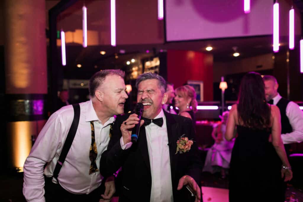 Two men singing into a microphone at a lively event with colorful lights. One man wears a tuxedo with a boutonniere, the other has a loosened tie and suspenders. People and decorations in background.