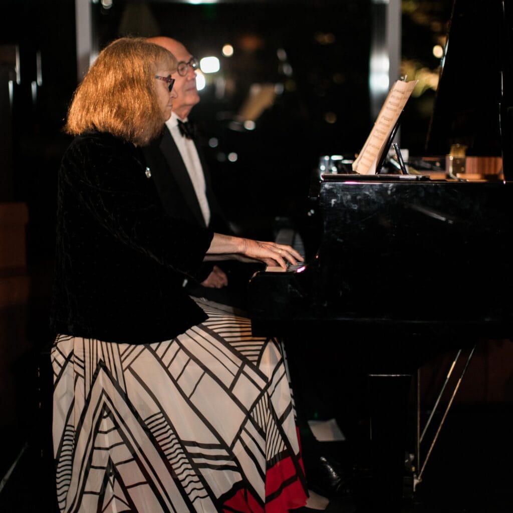 A woman and a man sit side by side at a piano, playing and reading sheet music together in a dimly lit setting—a quiet moment that hints at a love story with subtle, disco-inspired charm.