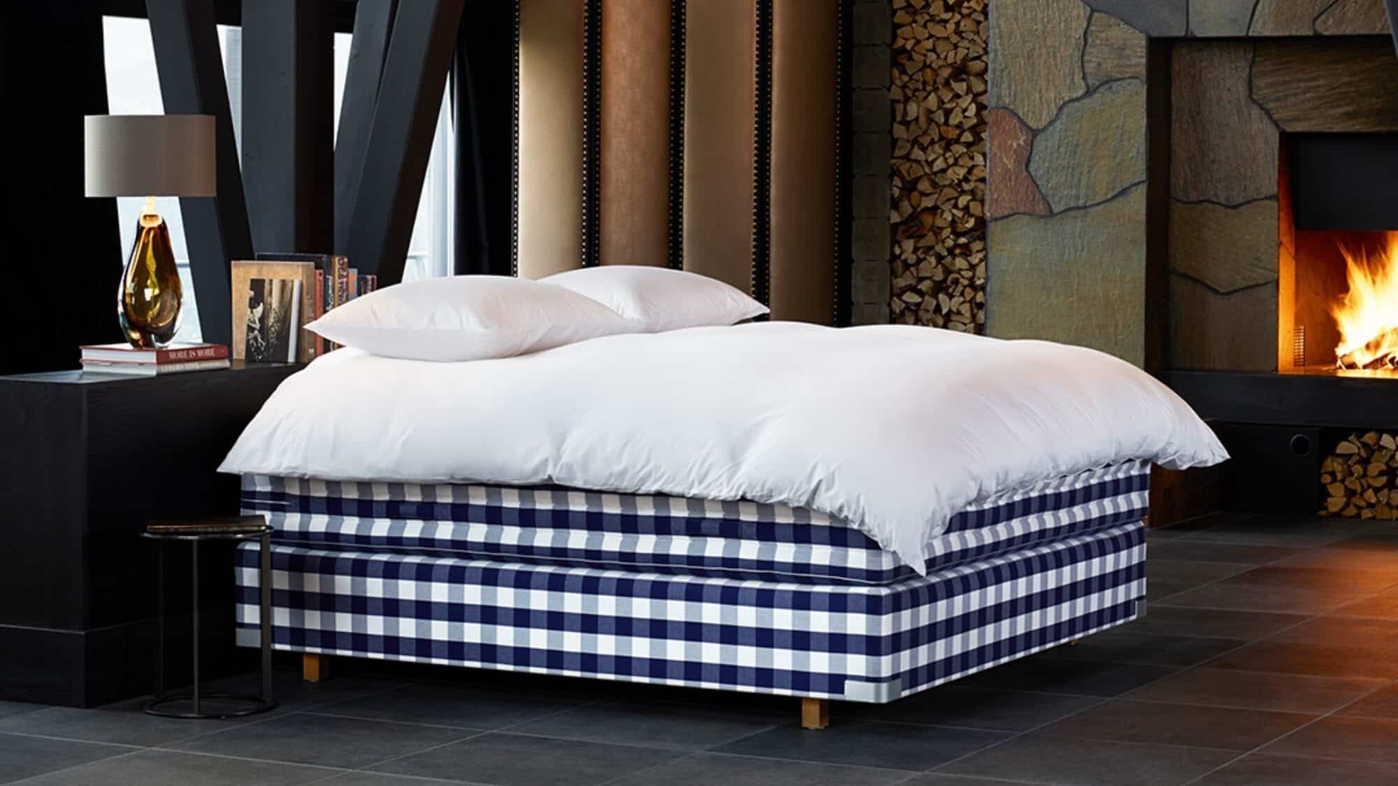 20130829_hastens_0246-16-9-2000x1125 A bed with white bedding and a blue plaid base in a modern room with a fireplace, books, and a lamp on a nightstand.