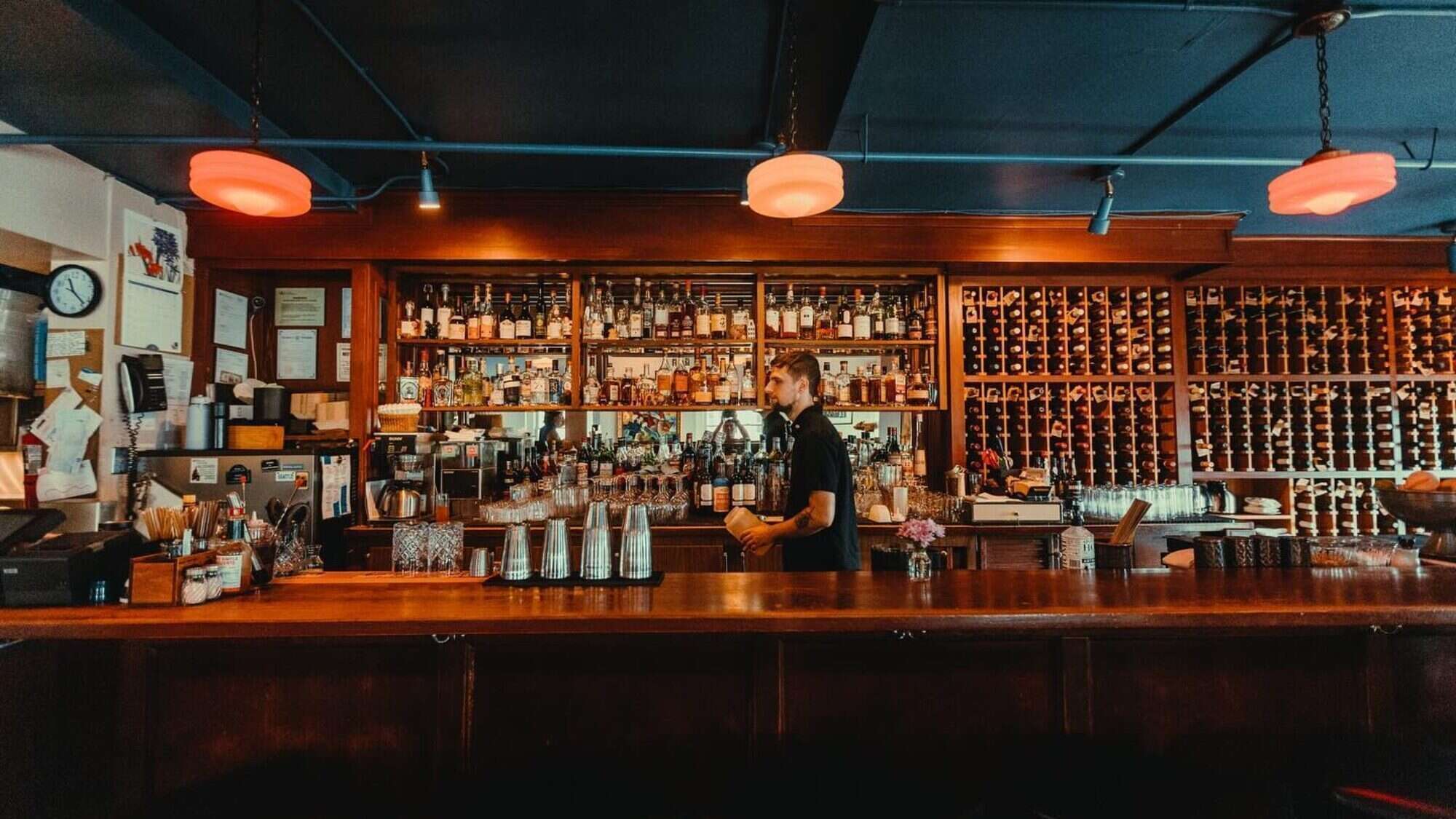 469546534_18472712359002219_907726171588578098_n-16-9-2000x1125 A bartender stands behind a wooden bar counter with shelves of liquor bottles and wine in the background, under warm ceiling lights.