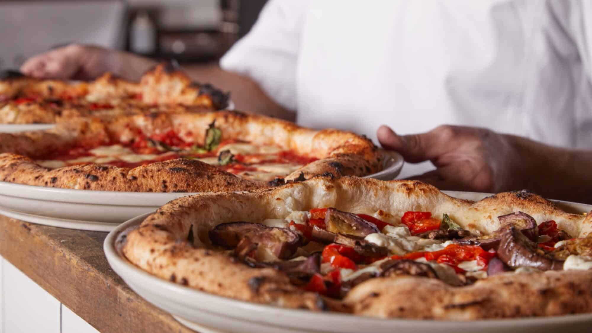 483576746_1111494800991684_3774367326140613776_n-16-9-2000x1125 Two freshly baked pizzas with various toppings are served on white plates by a person in a white shirt.