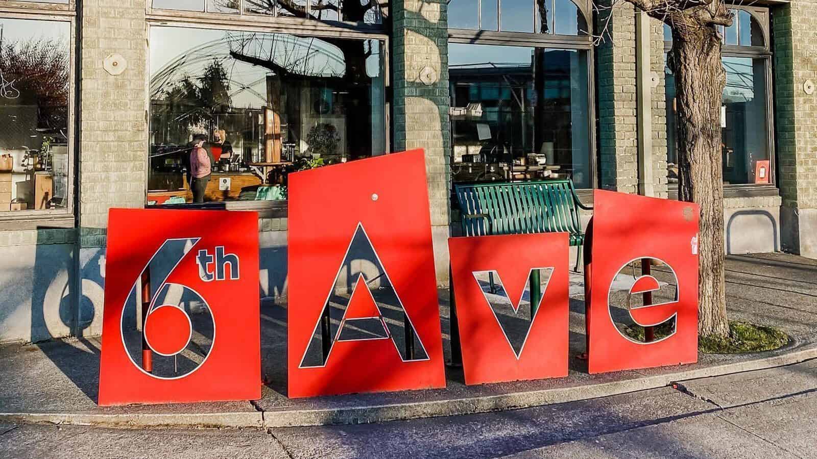 Large red signs spelling "6th Ave" are displayed on a sidewalk in front of a brick building, its large windows reflecting the street and trees—capturing the vibrant spirit of Heart of Glass: A Weekend in Tacoma.
