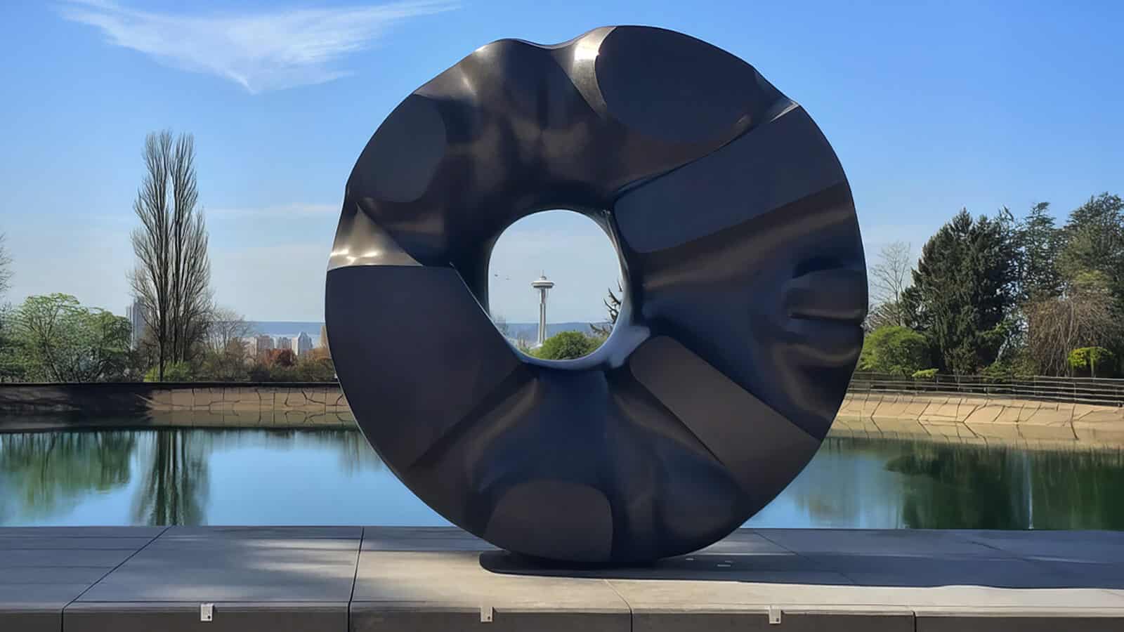 BlackSun-Seattle_PeterAlfredHess-Flickr_1600x900 Large black abstract circular sculpture with a central hole, displayed outdoors near a reflective pool, stands among trees and a tower—one of the striking Seattle cultural landmarks.