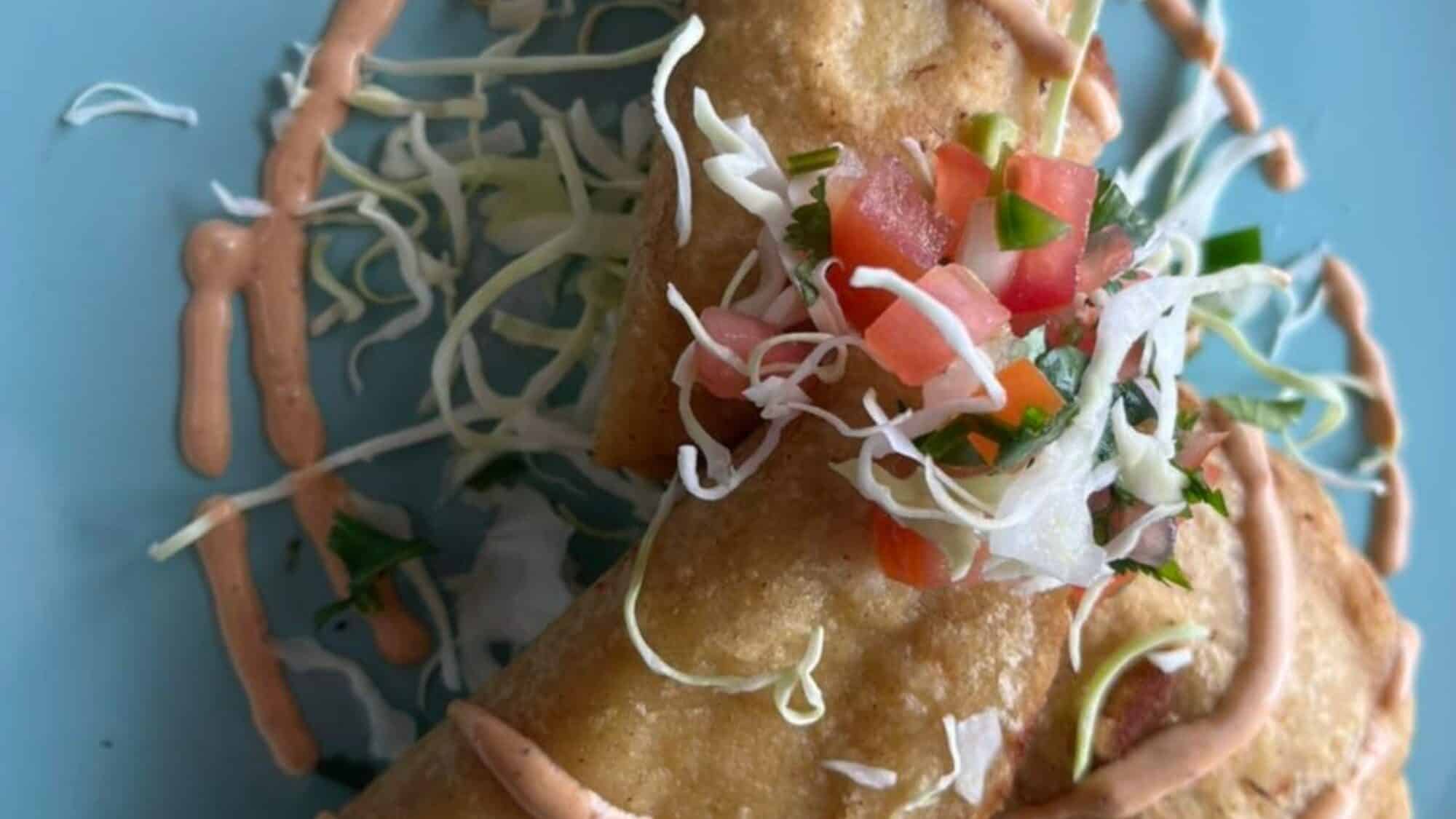 Blue crab empanadas are one of the specialties at El Cabrito in Burien-16-9-2000x1125 Two fried tacos topped with shredded cabbage, diced tomatoes, onions, and cilantro, drizzled with a creamy sauce, served on a blue plate.