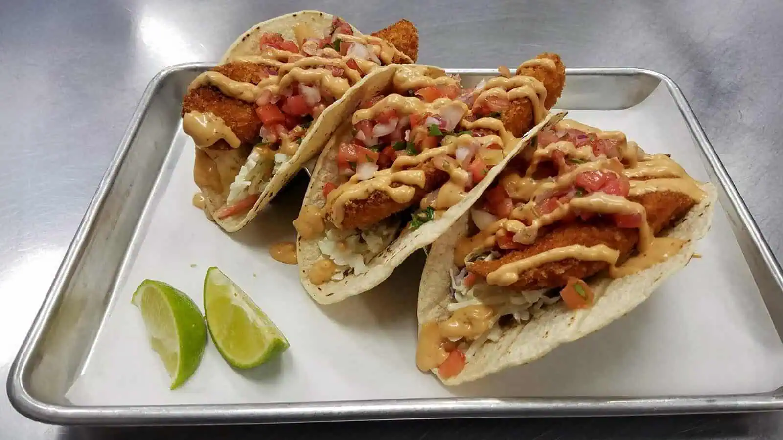 BurienFishHouse-FishTacoTrio_1600x900 Three breaded fish tacos from Counter Culture: Burien Fish House, topped with diced tomatoes, shredded cabbage, and creamy sauce on tortillas, served on a metal tray with two lime wedges.