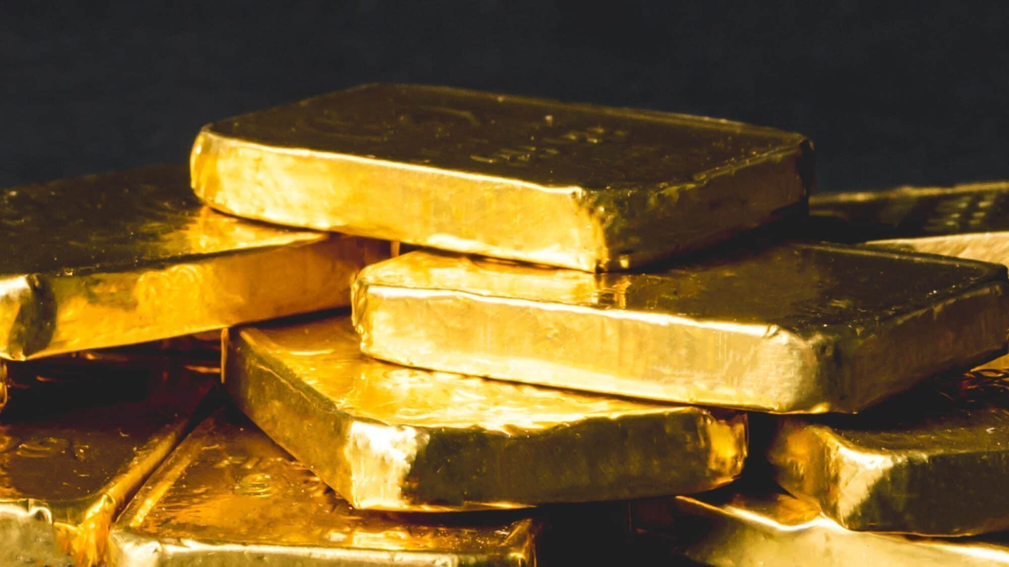 COSTCO GOLD-16-9-2000x1125 Several shiny gold bars are stacked unevenly against a dark background.