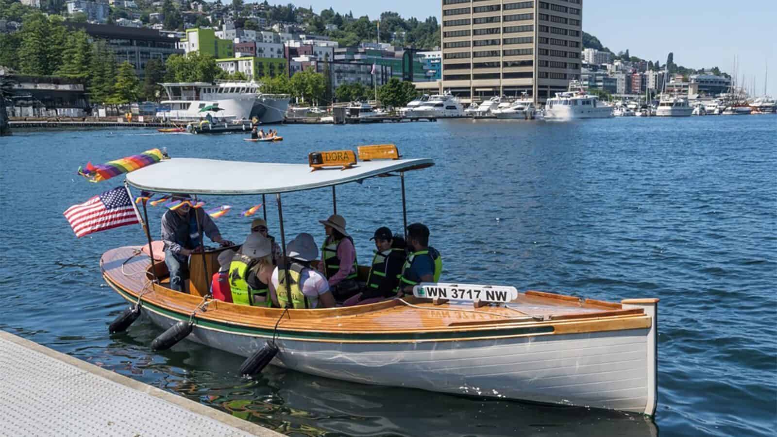 A small electric boat with passengers in life jackets is docked on a lake during Seattle summer, with city buildings and other boats in the background.