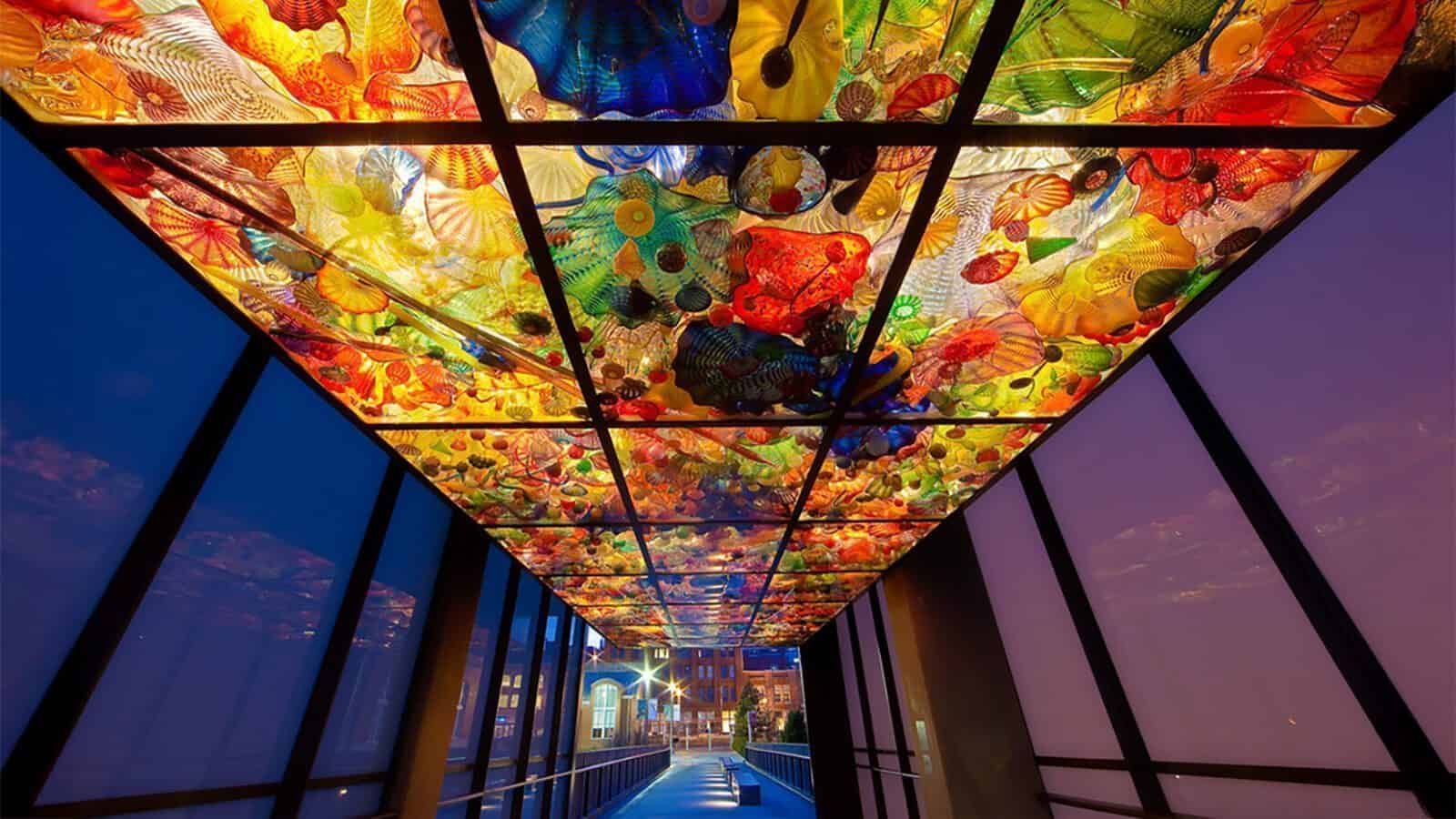 A walkway with glass walls features a vibrant, colorful glass artwork ceiling illuminated from above, displaying abstract shapes and patterns—an experience reminiscent of Heart of Glass: A Weekend in Tacoma.