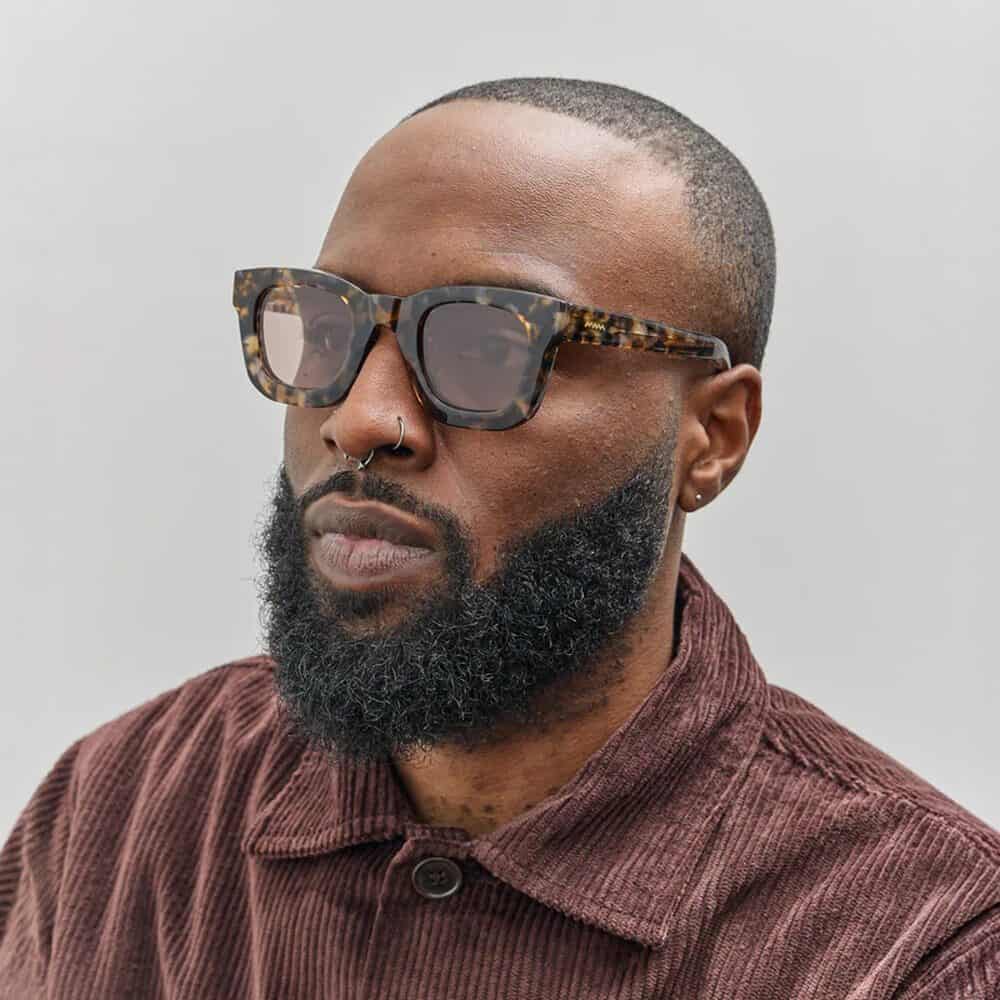 A man with a full beard and mustache wears tortoiseshell sunglasses, a nose ring, and a brown corduroy jacket against a plain light gray background—perfect inspiration for your Seattle Father's Day gifts.