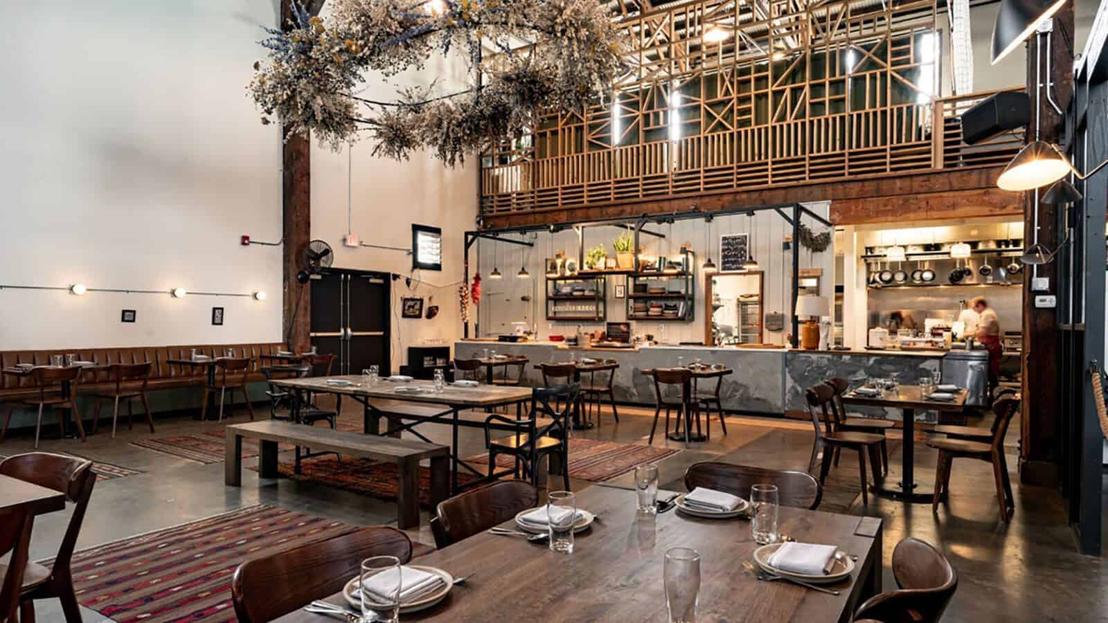 Spacious restaurant interior with wooden tables and chairs, modern decor, open kitchen, high ceiling, and hanging dried floral arrangement inspired by Oregon wine country.