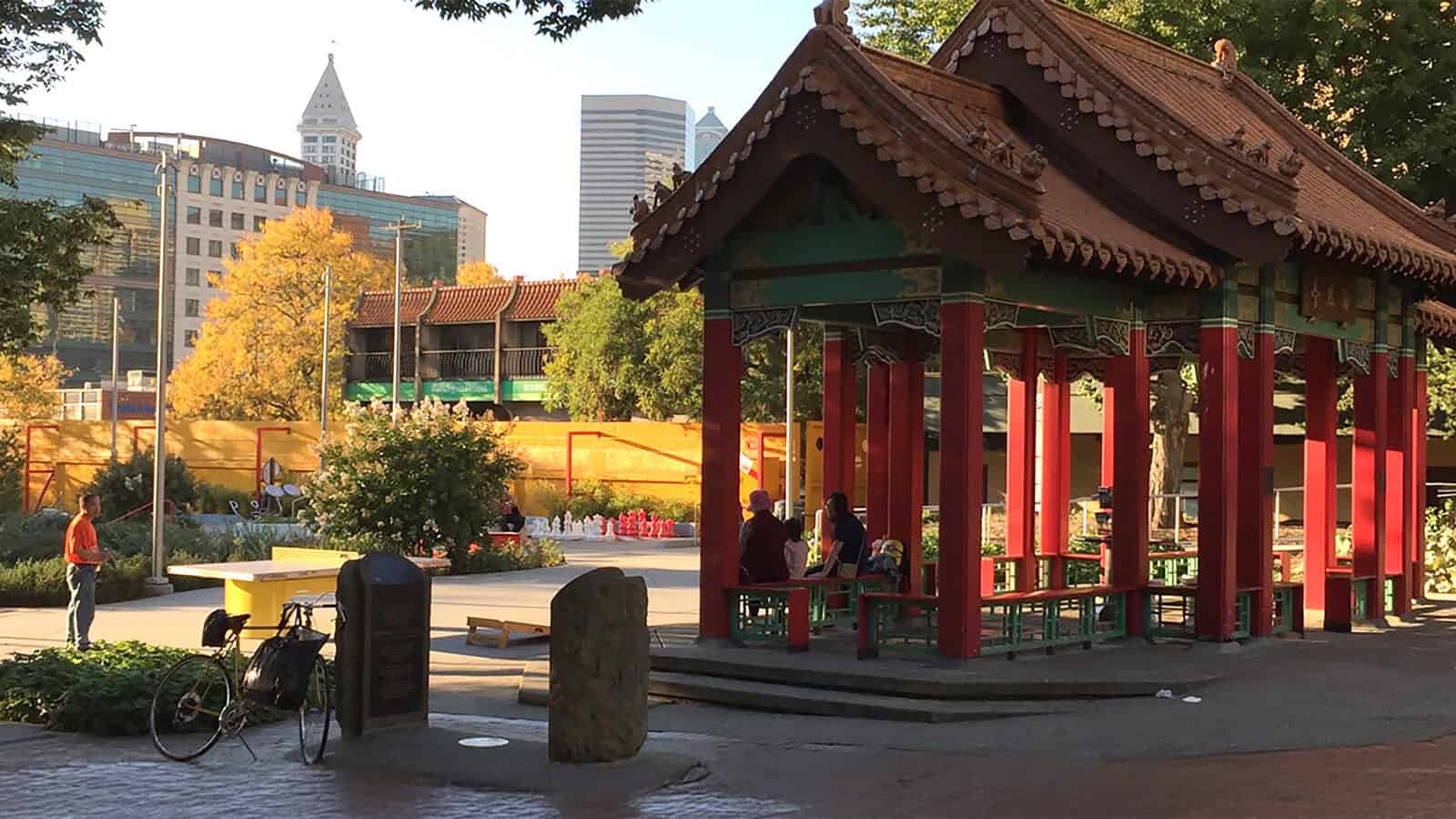 A traditional Chinese pavilion stands in a Seattle city park with a few people sitting inside; modern buildings and trees frame this cultural landmark, cherished by the AANHPI community.